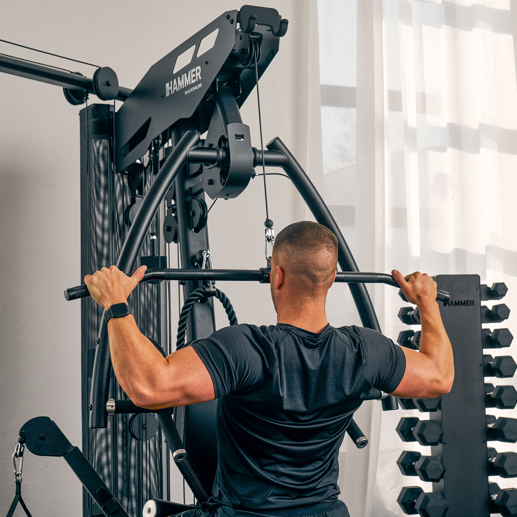 Mann trainiert an einer schwarzen Fitnessmaschine, abgewandt. Er trägt schwarze Kleidung. Ein Gewichtsregal befindet sich hinter ihm.