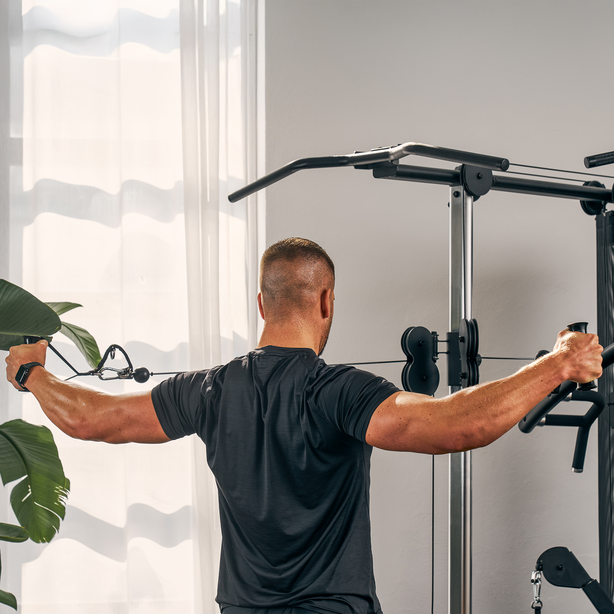 Ein Mann trainiert an einem Fitnessgerät, Arme ausgestreckt, Rücken zur Kamera, vor einem Fenster und einer Pflanze.