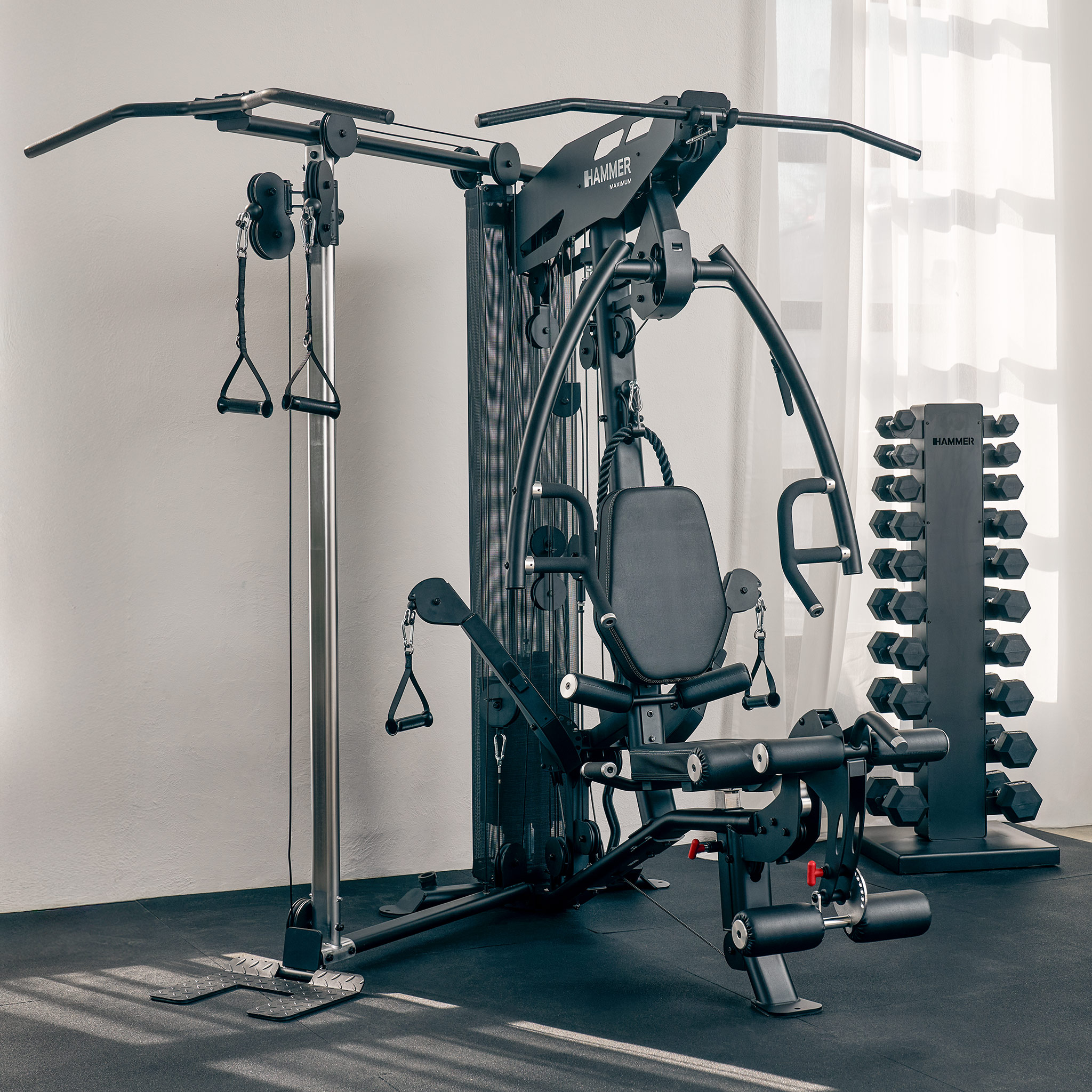 Eine große schwarze Fitnessmaschine mit verschiedenen Trainingsstationen. Rechts steht ein Hantelständer, in der Nähe eines Fensters.