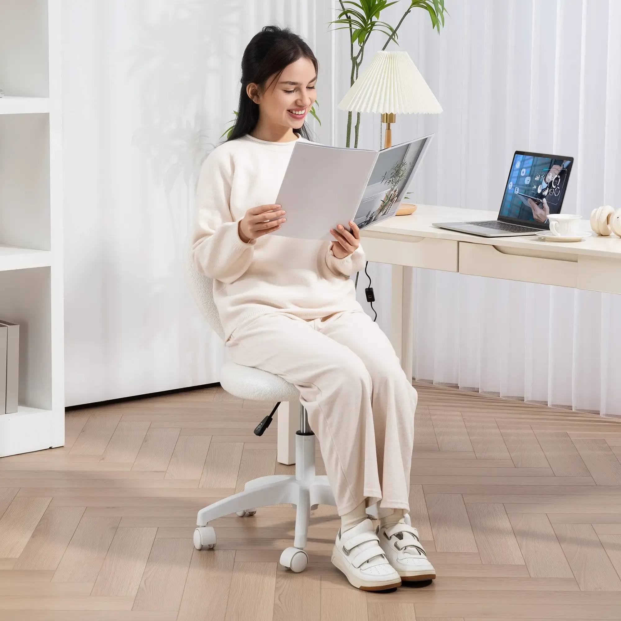 Frau in weißem Outfit liest eine Zeitschrift, sitzt auf einem weißen Stuhl vor einem Schreibtisch mit Laptop.