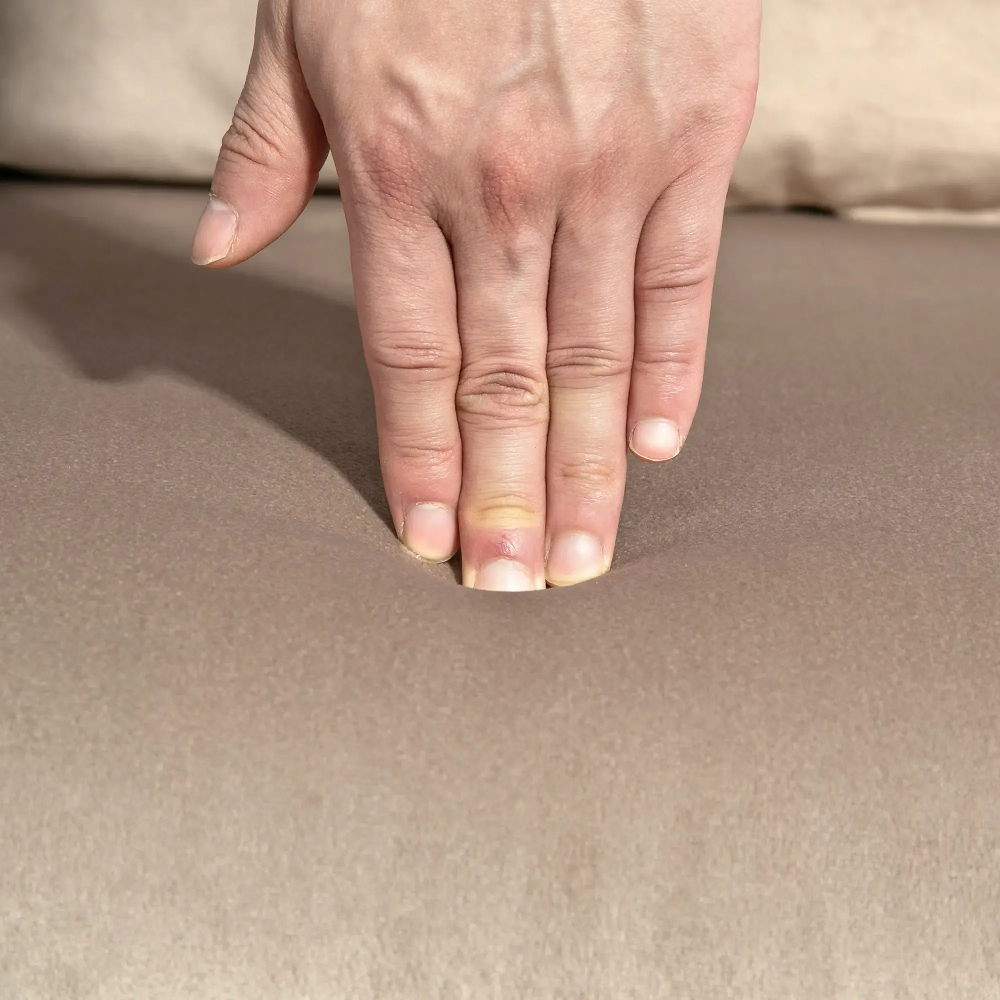 Eine Hand drückt in ein hellbraunes Kissen. Der Mittelfinger hat eine rote, geschwollene Stelle.