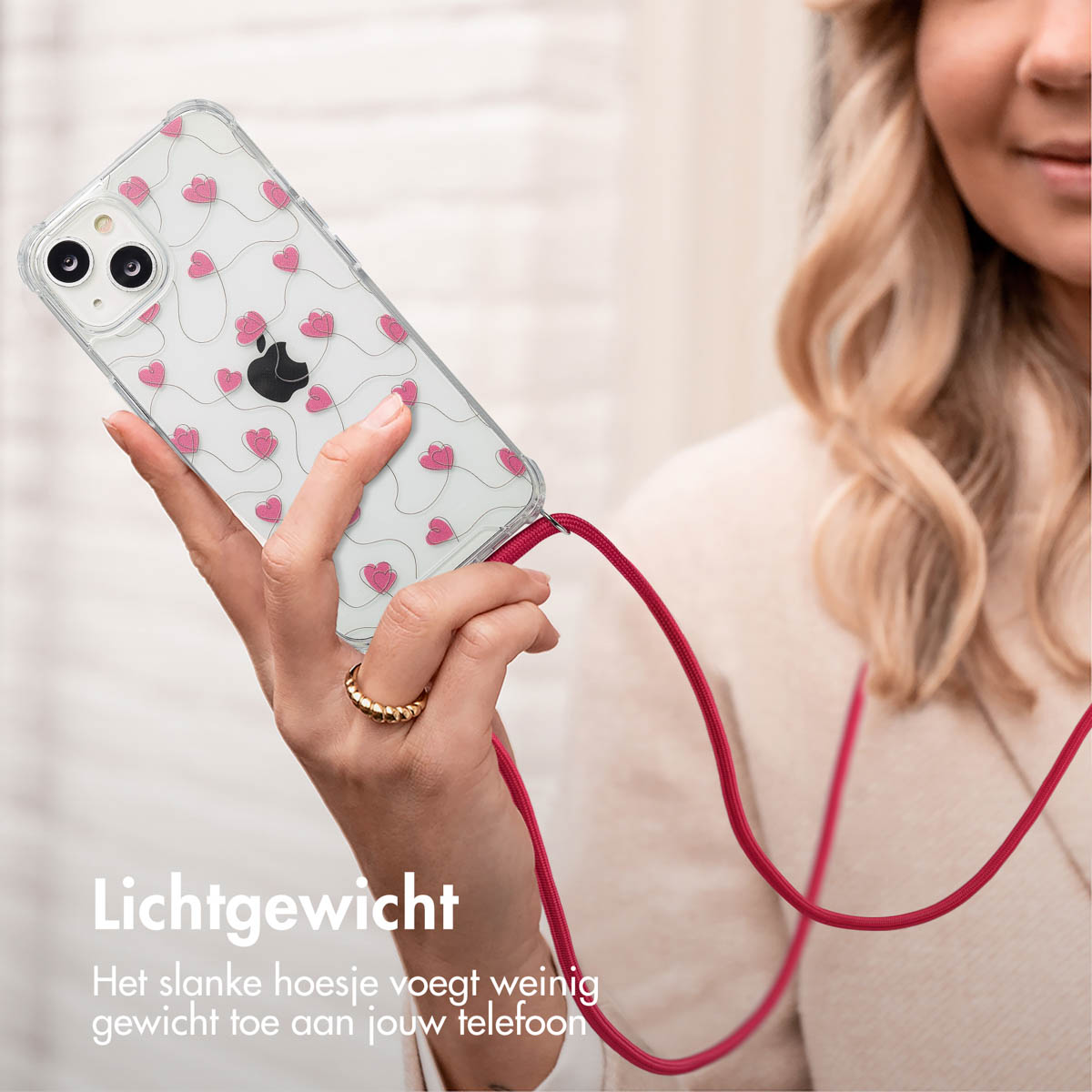 Een vrouw houdt een telefoon vast met een hoesje met hartjespatroon en een rood koord. Ze draagt een ring.
