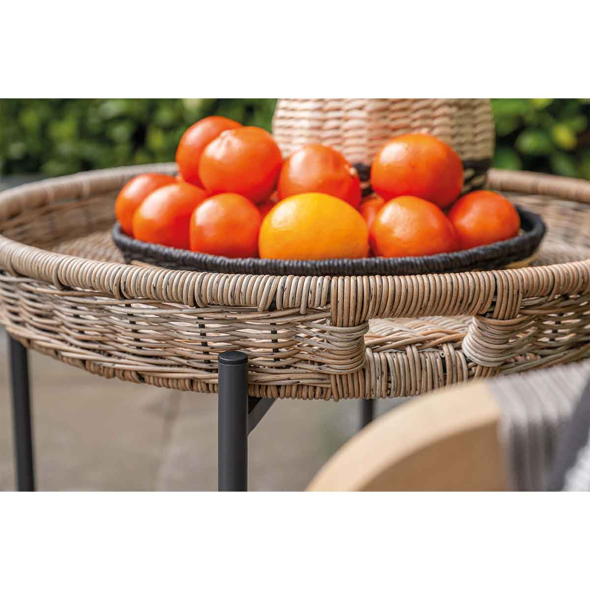 A wicker tray with tangerines on a table, outdoors, with a black stand and a green background.