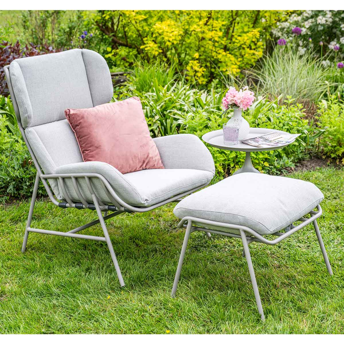 A gray chair with a pink pillow and a small table in a garden setting. Flowers are on the table.