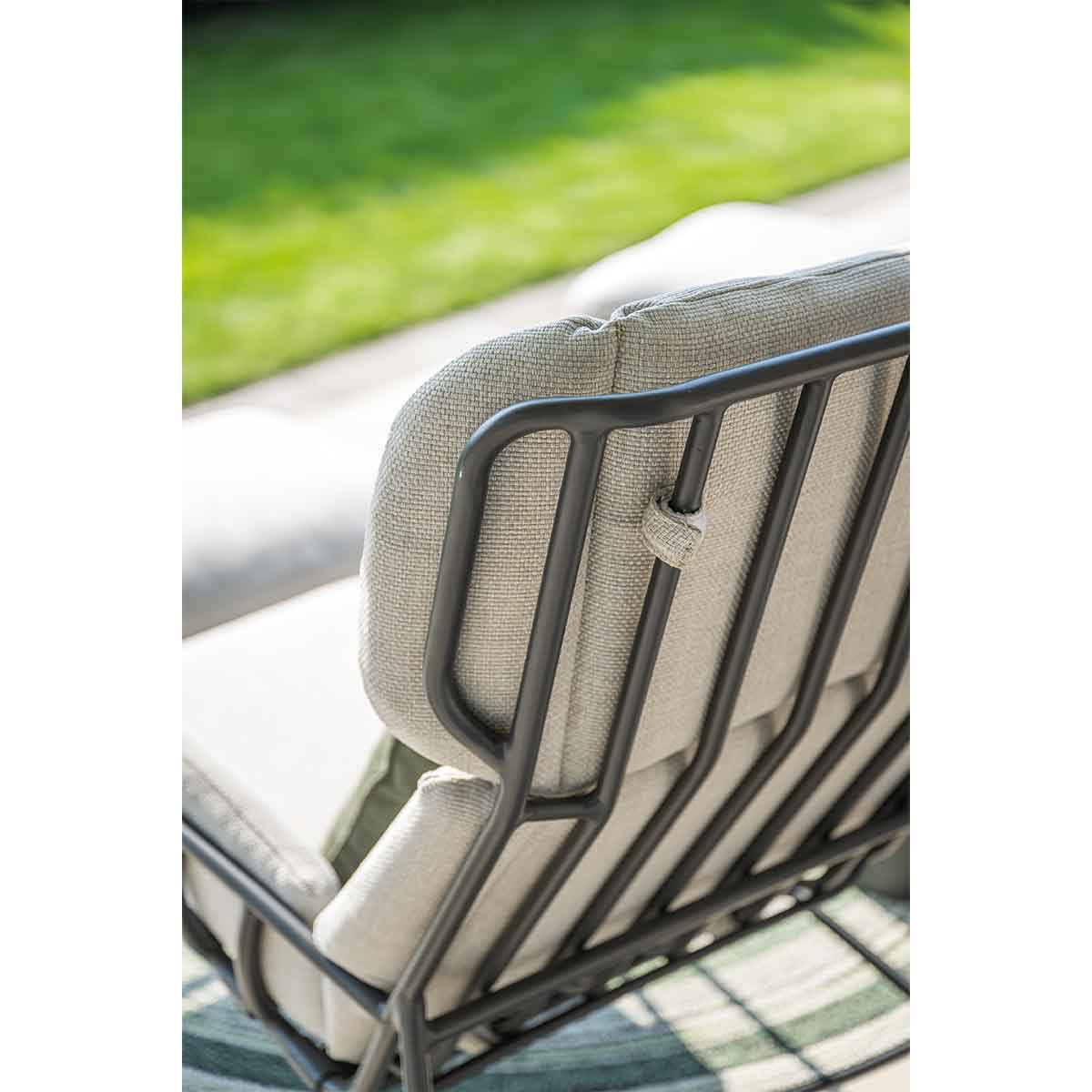 Close-up of a chair's backrest. It has a dark frame and light gray cushion. Set on a green and white patterned rug.