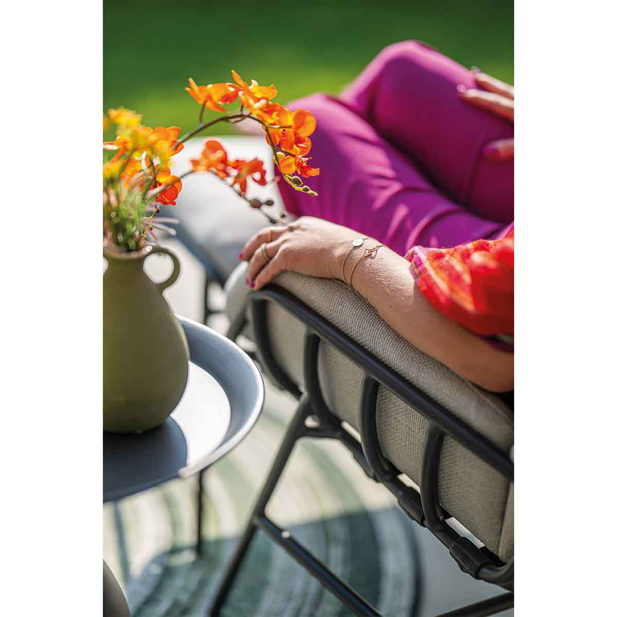 A person is sitting in a chair, arms resting, next to a table with a vase of flowers. The person is wearing pink.