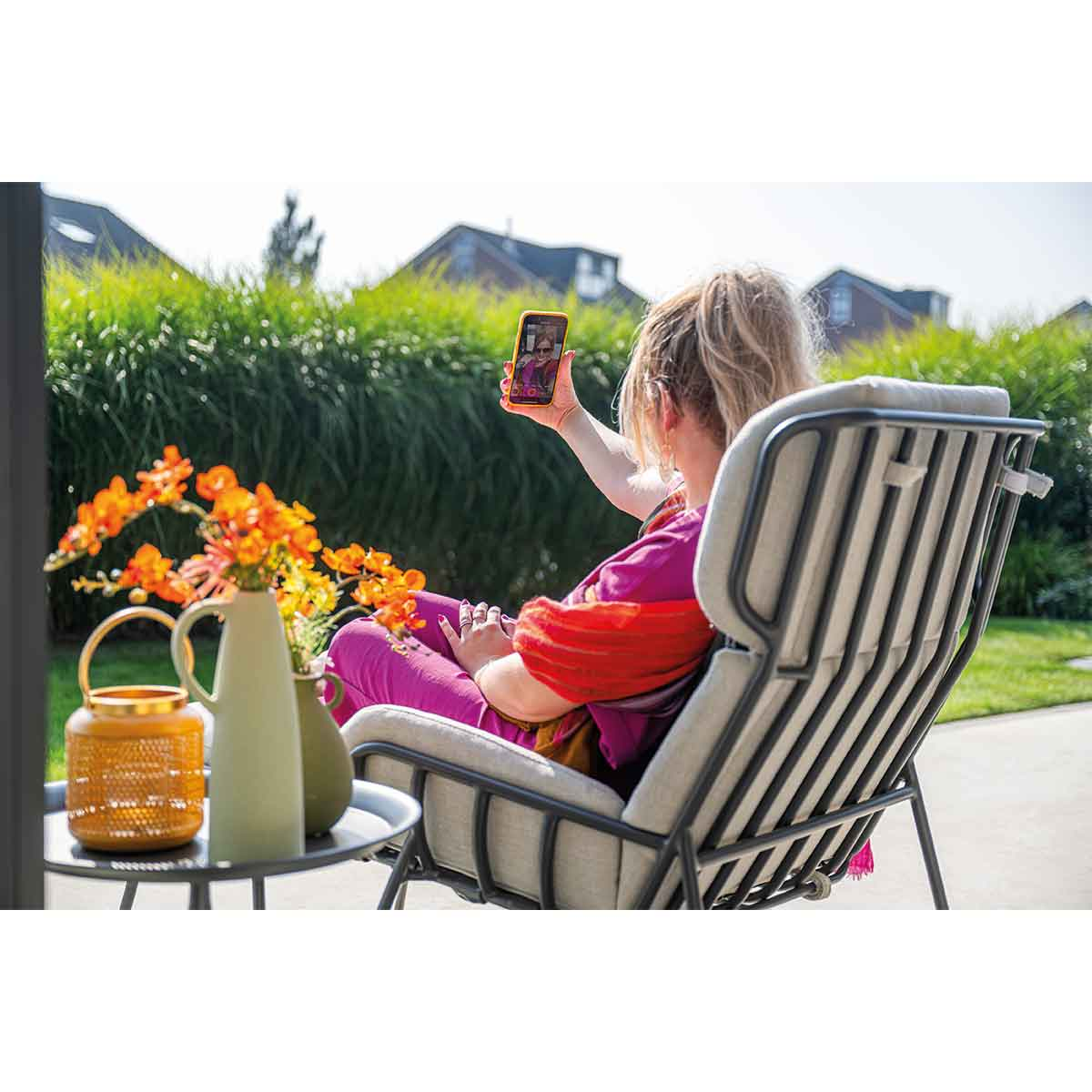 A woman is sitting in a gray chair in a garden and taking a selfie with her phone. Flowers and table are present.