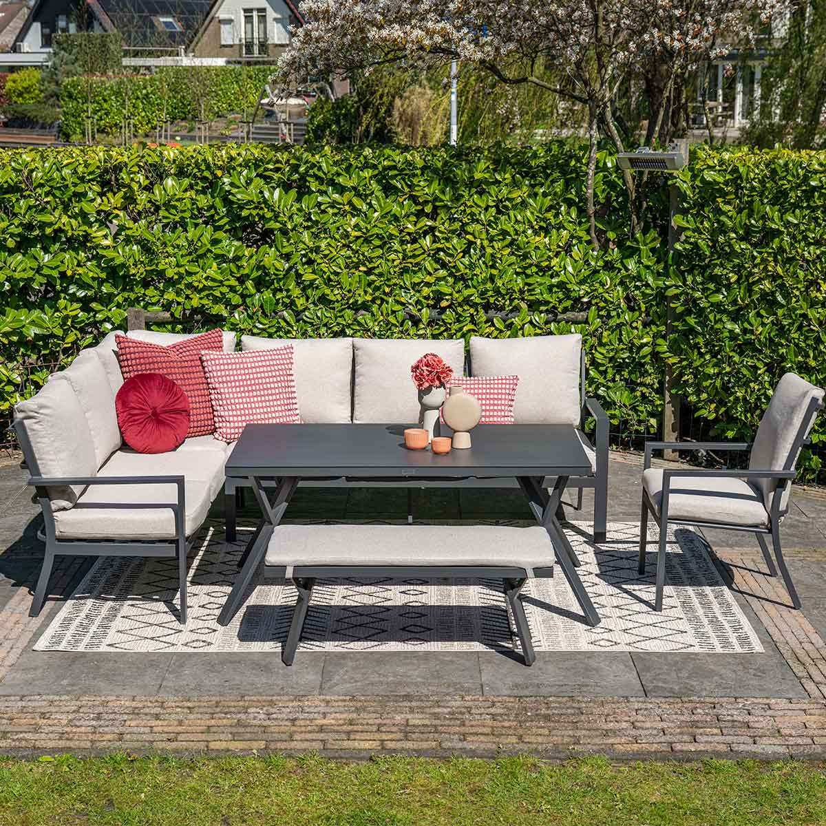Outdoor seating set with gray table, bench, and chairs on a patterned rug, green hedge background.