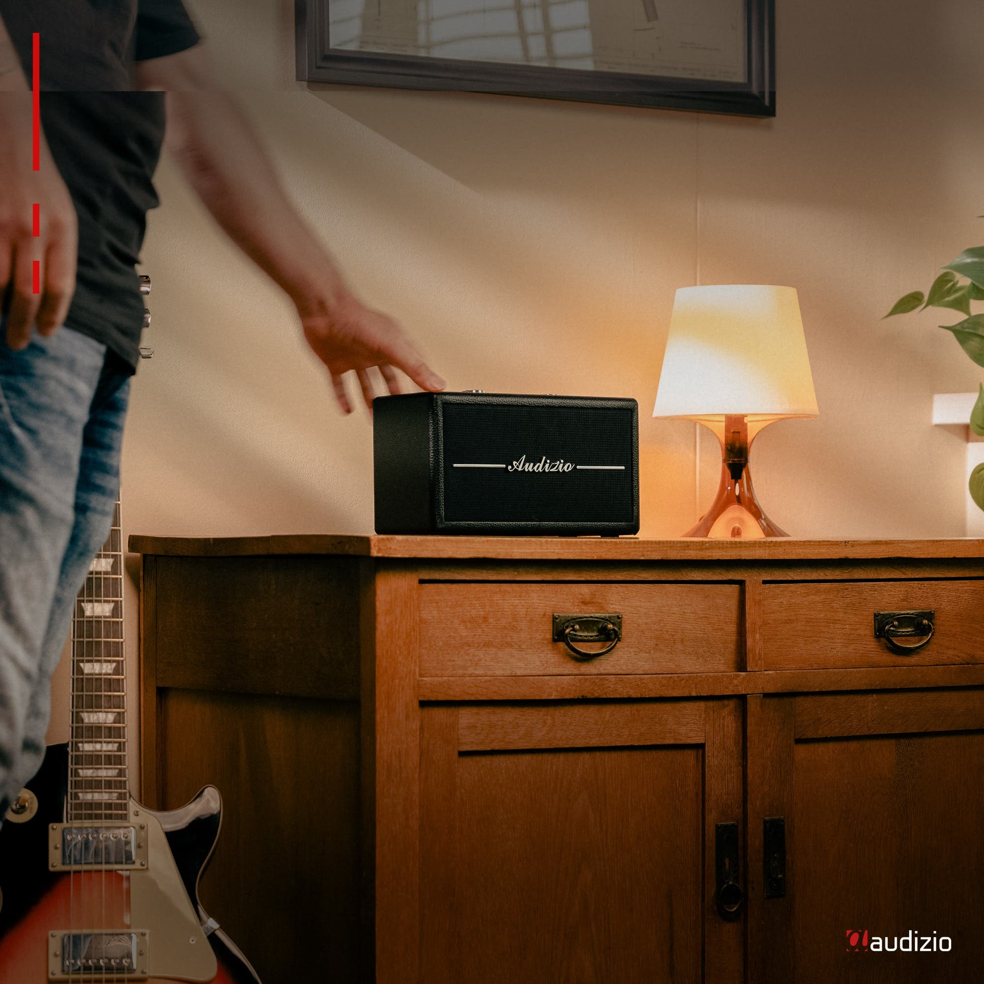 Mano de una persona alcanzando un altavoz negro en un mueble de madera. Hay una guitarra a la izquierda.