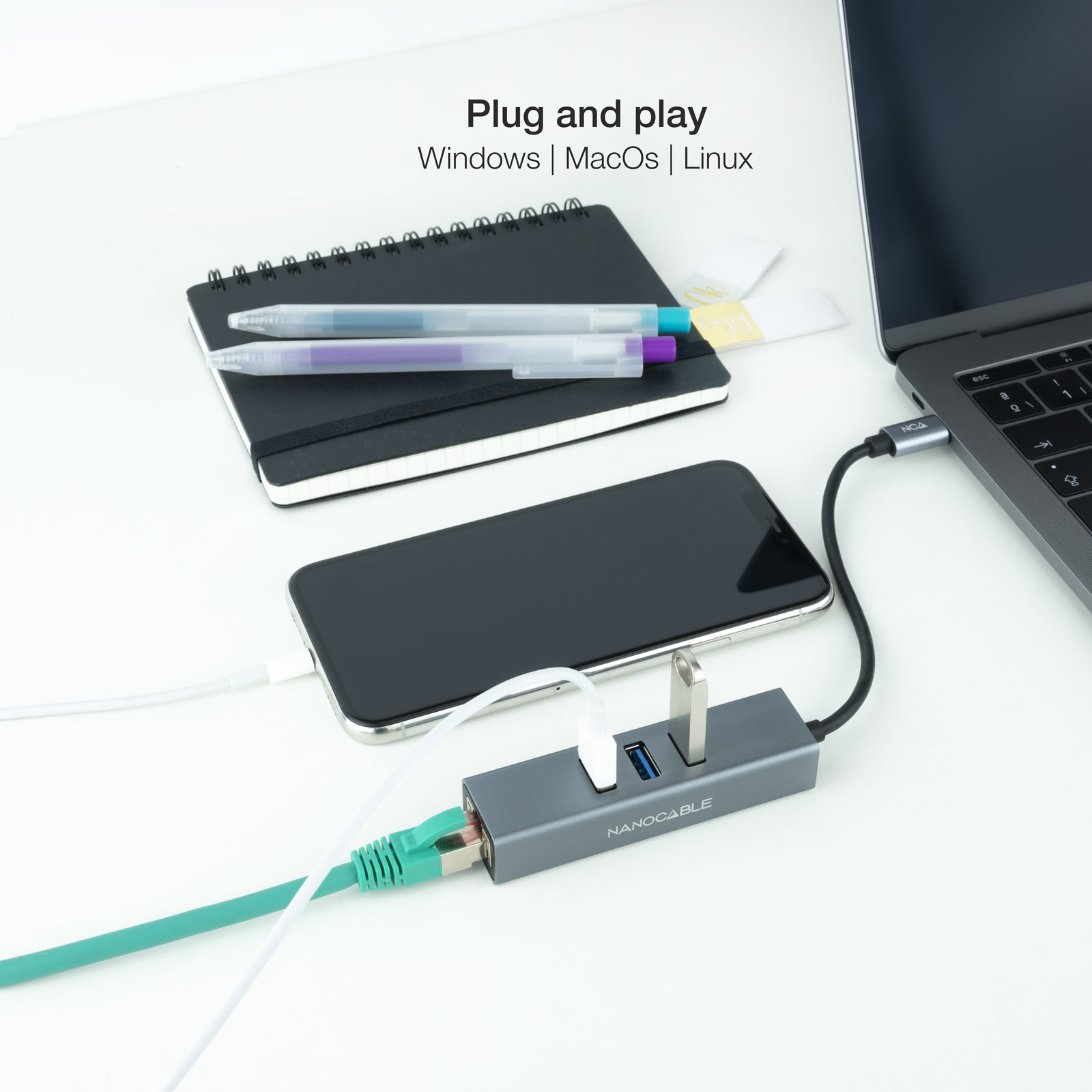 Laptop conectado a un hub, teléfono, bolígrafos y cuaderno sobre superficie blanca.