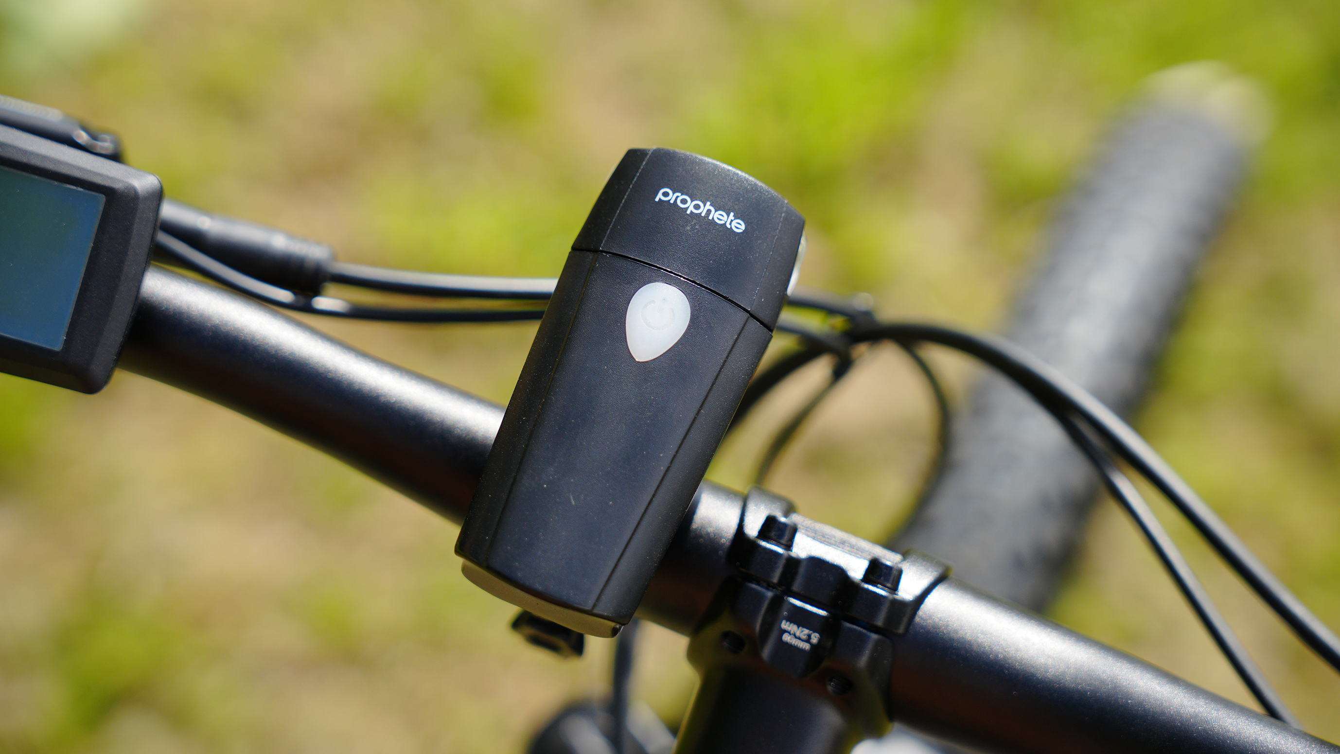 Eine schwarze Prophete Fahrradlampe mit einem weißen Knopf ist an einem schwarzen Lenker mit grünem Hintergrund montiert.