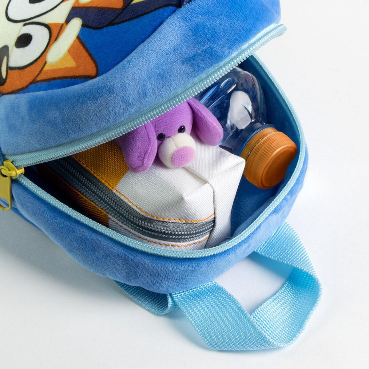 Mochila azul con cremallera abierta, con un juguete morado, una caja blanca y una botella de agua.