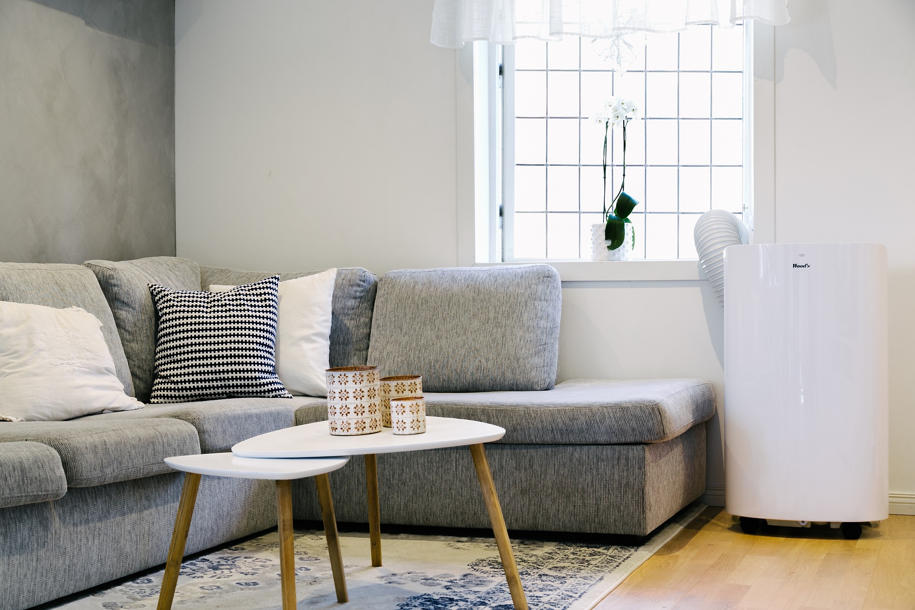 Wohnzimmer mit grauem Sofa, weißen Tischen und weißem Klimagerät am Fenster.