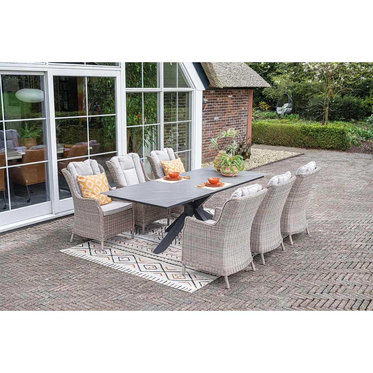 A patio setting with a long table, wicker chairs, and a stone floor. Houses in the background.
