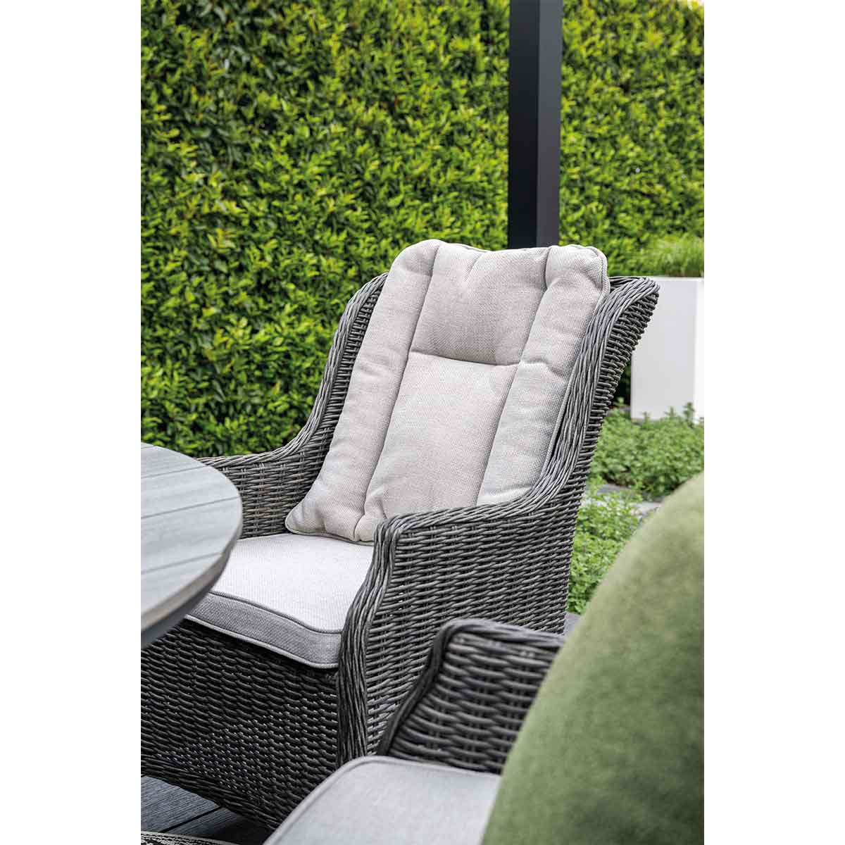 Close-up of a wicker chair with a gray cushion and a green hedge in the background.
