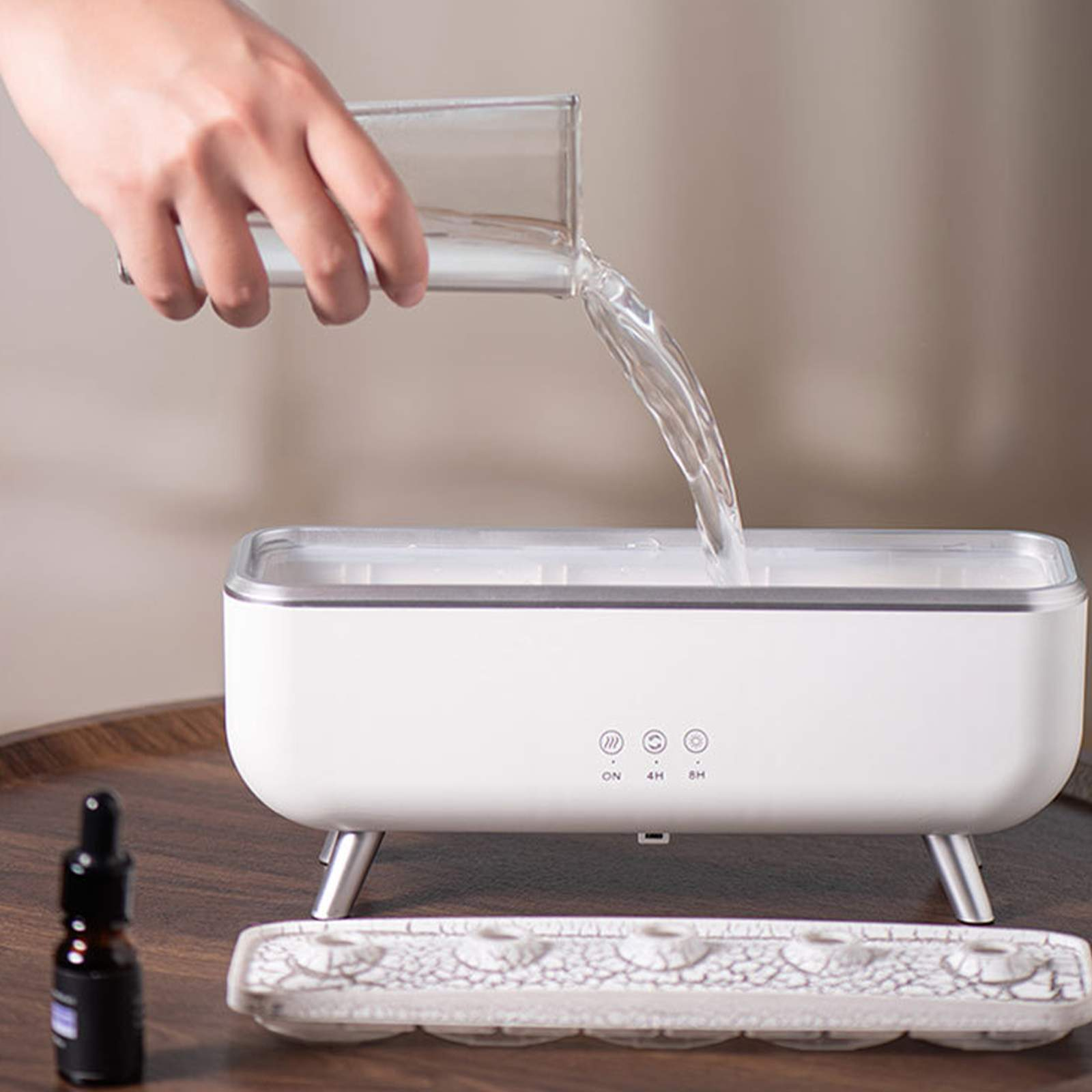 Hand gießt Wasser in weißen Luftbefeuchter. Kleine Schalen und Ölflasche daneben.
