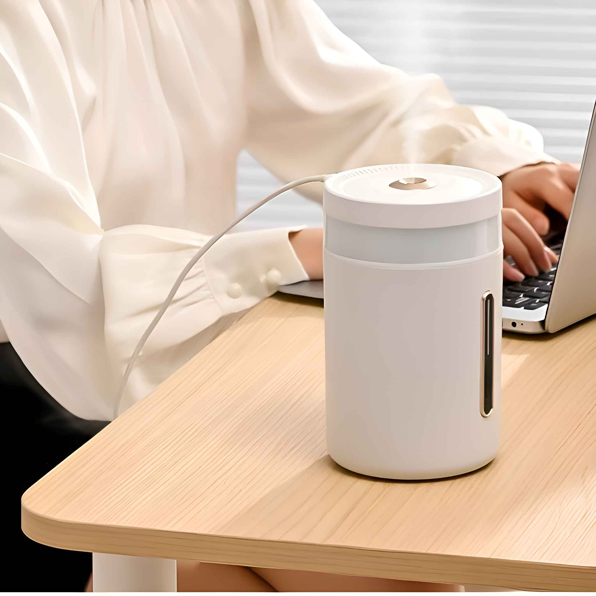 Ein weißer Luftbefeuchter steht auf einem Holzschreibtisch. Eine Person tippt im Hintergrund auf einem Laptop.