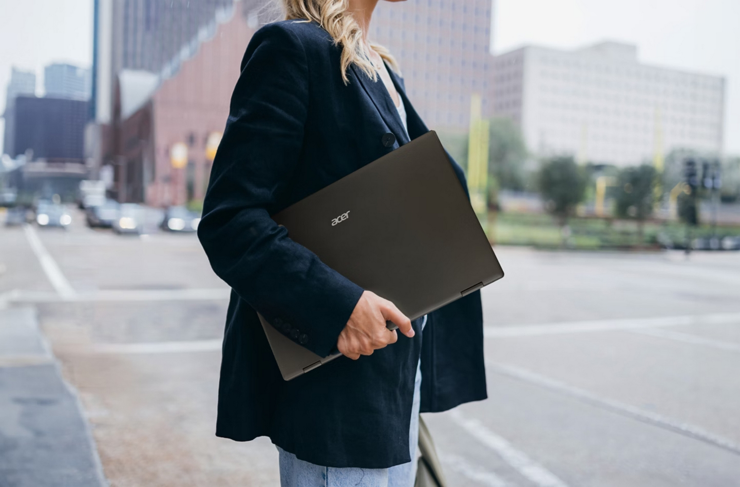 Kobieta w marynarce trzyma zamknięty laptop. Ciemny laptop, logo Acer. Tło miejskie.