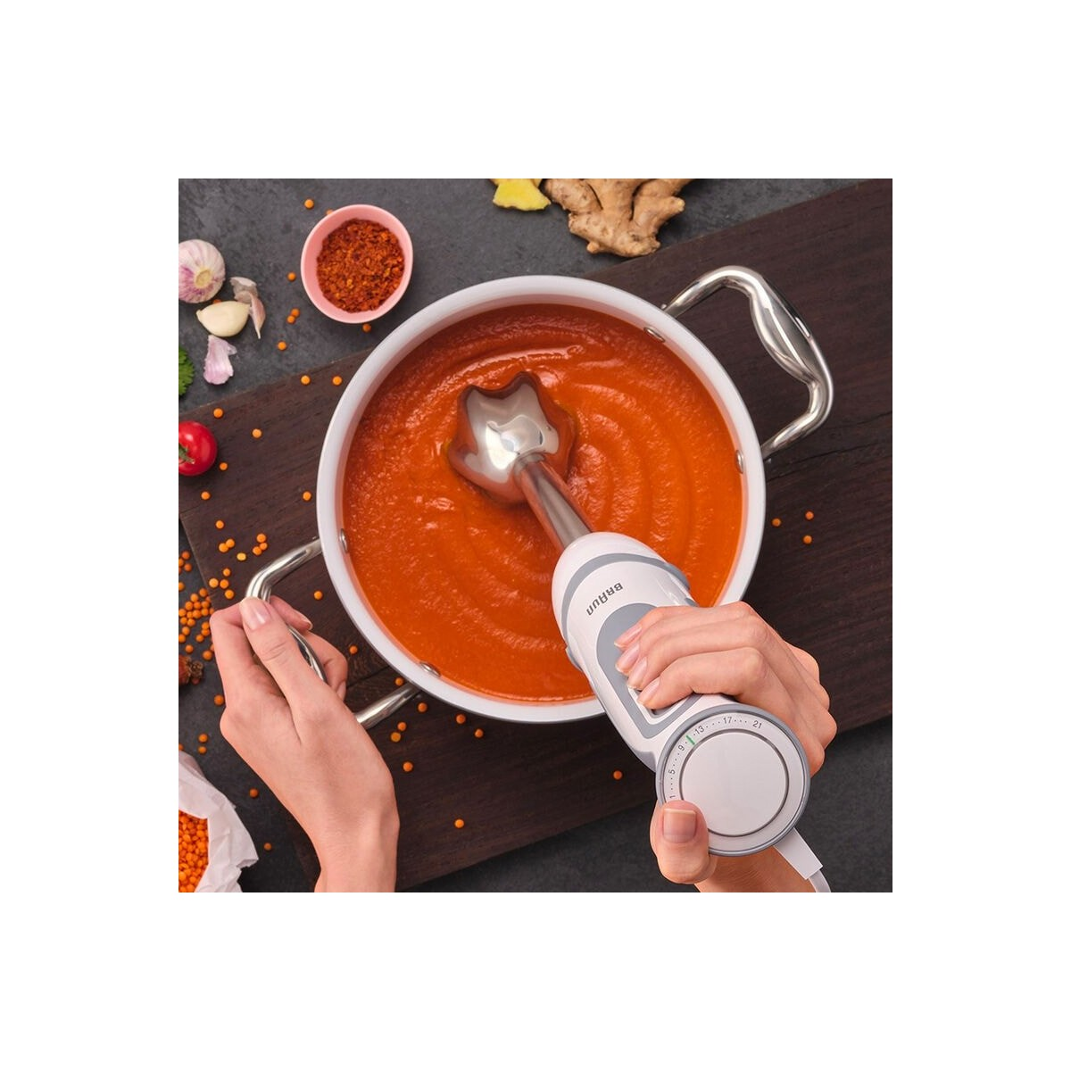 Una mano usa una batidora de mano en una olla de sopa naranja. Ingredientes alrededor de la olla.