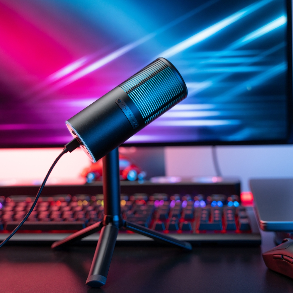 Un microfono nero su un supporto, illuminato con luci blu e rosa. Una tastiera e un monitor sullo sfondo.