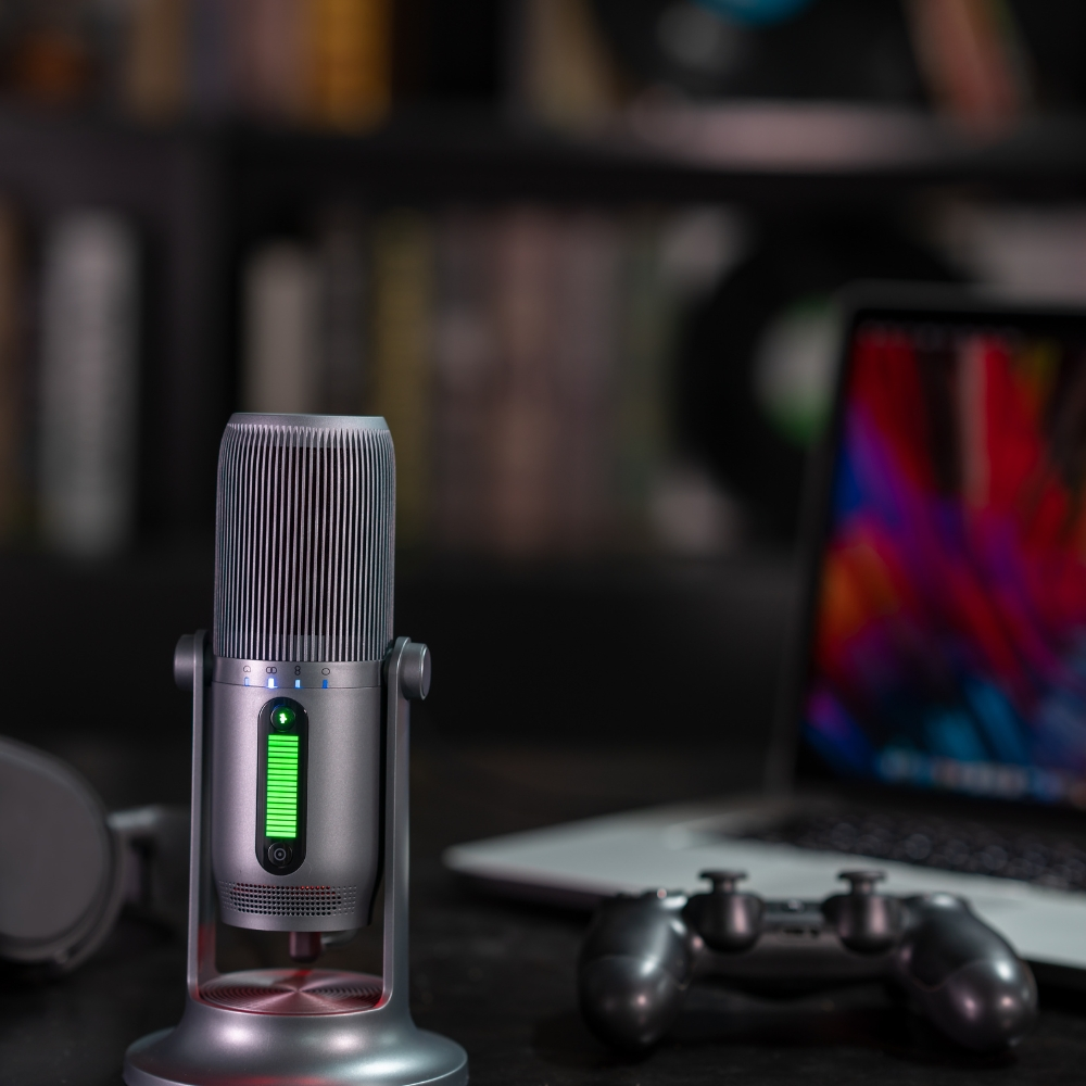 Microfono argento con luci verdi. Un laptop, un controller e delle cuffie sullo sfondo sfocato.