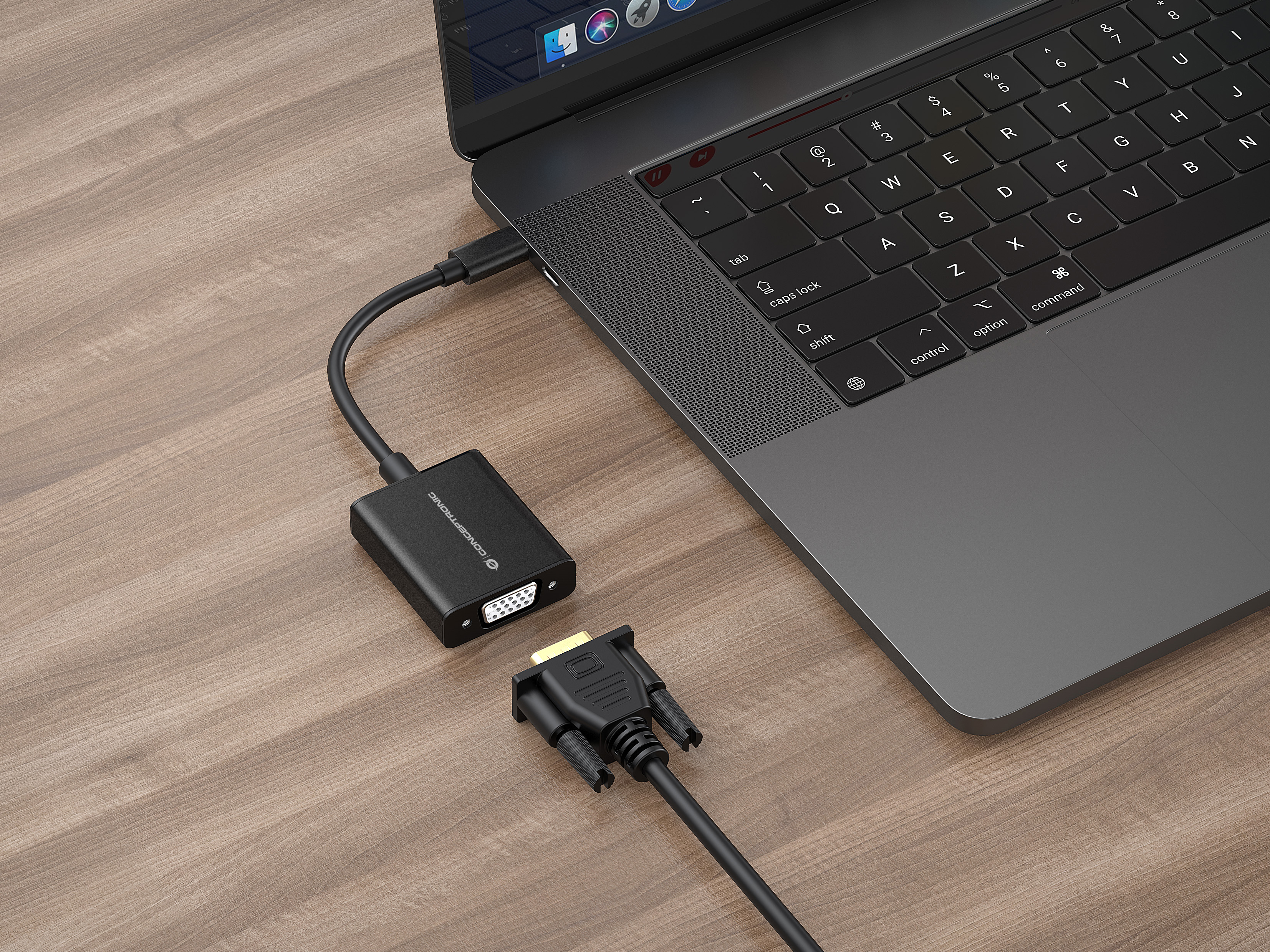 Un adaptador VGA Conceptronic está conectado a un portátil sobre una superficie de madera, con otro cable VGA cerca.