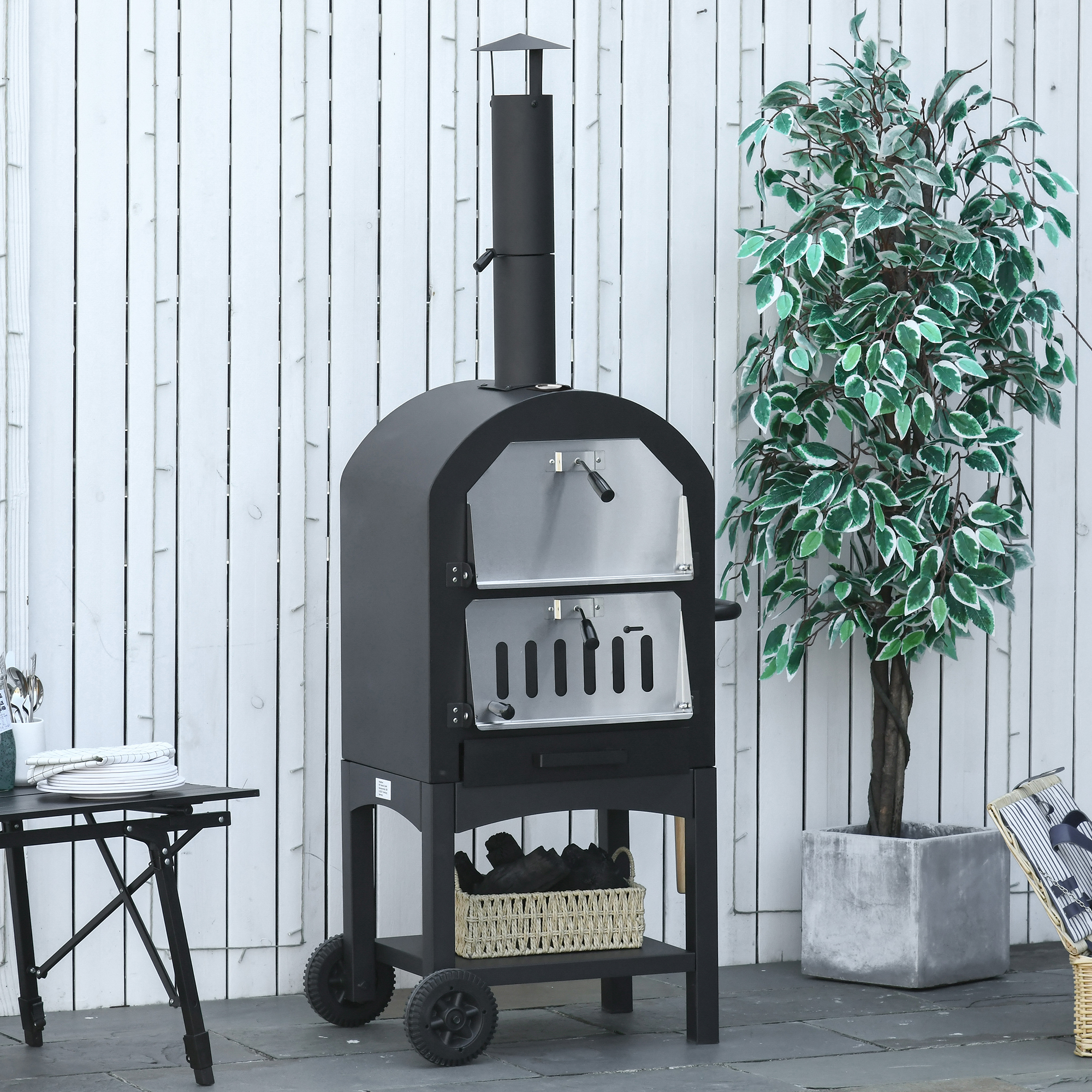 Un forno per pizza nero da esterno con camino, accanto a una pianta, un tavolo e una recinzione bianca. Sotto c'è un cesto di carbone.