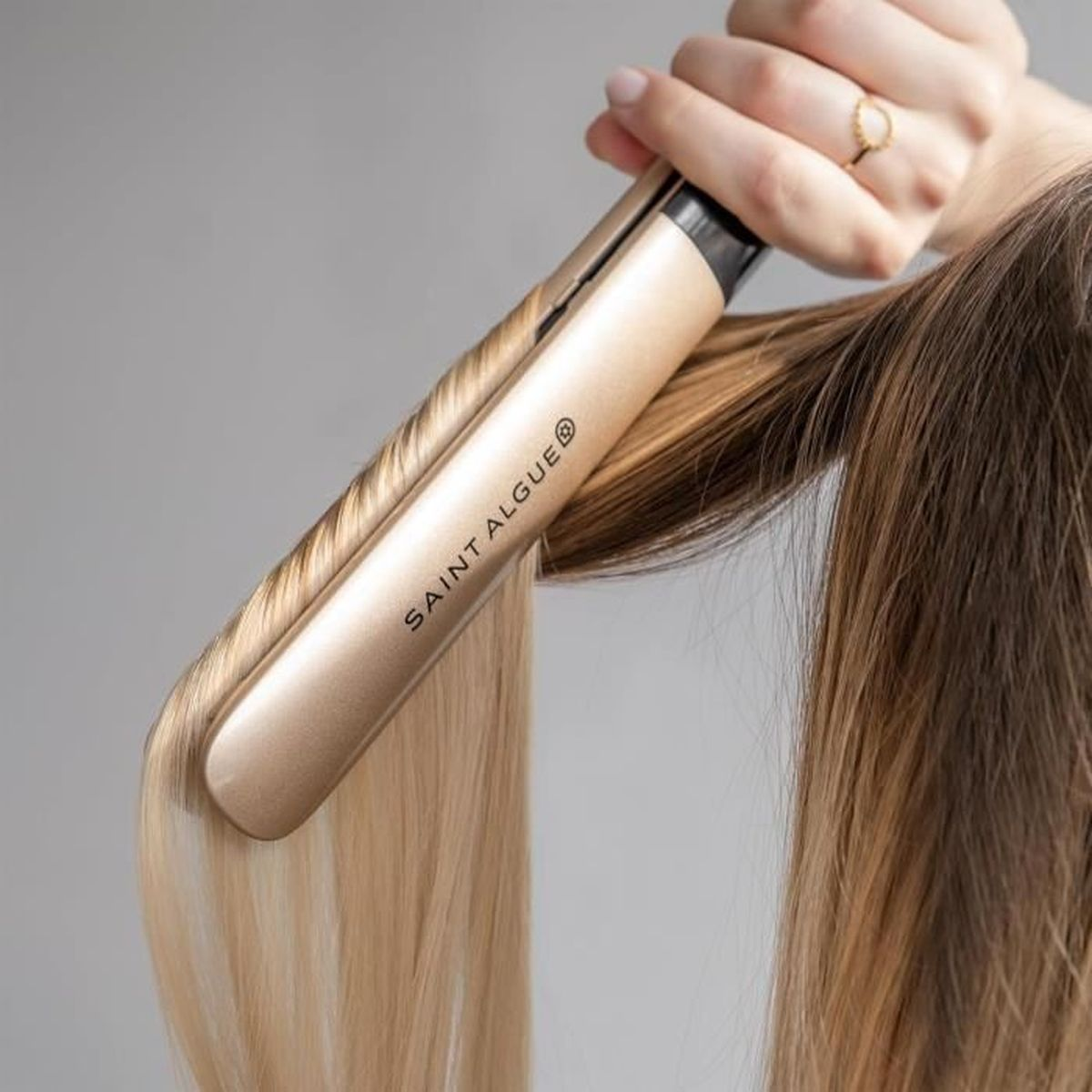 Eine Person, die blondes Haar mit einem goldfarbenen Saint Algue Haarglätter glättet.