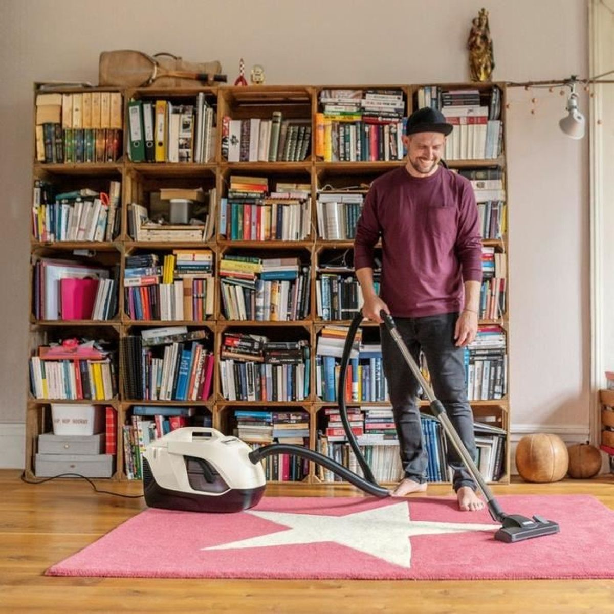 Mężczyzna odkurza różowy dywan ze gwiazdą przed regałem. Regały z drewnianych skrzyń.