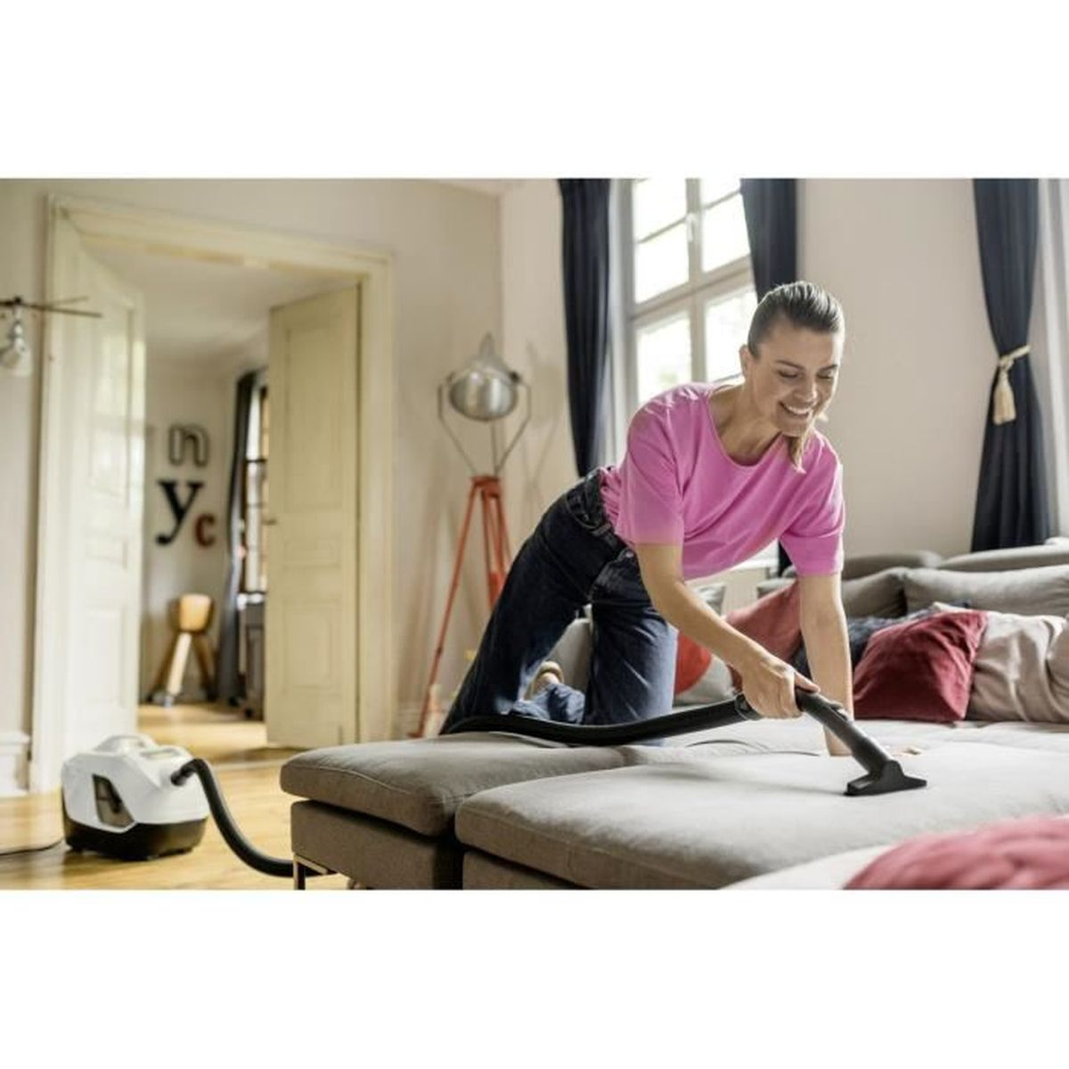 Kobieta odkurza sofę. Odkurzacz po lewej. Wnętrze ze sztuką na ścianie.