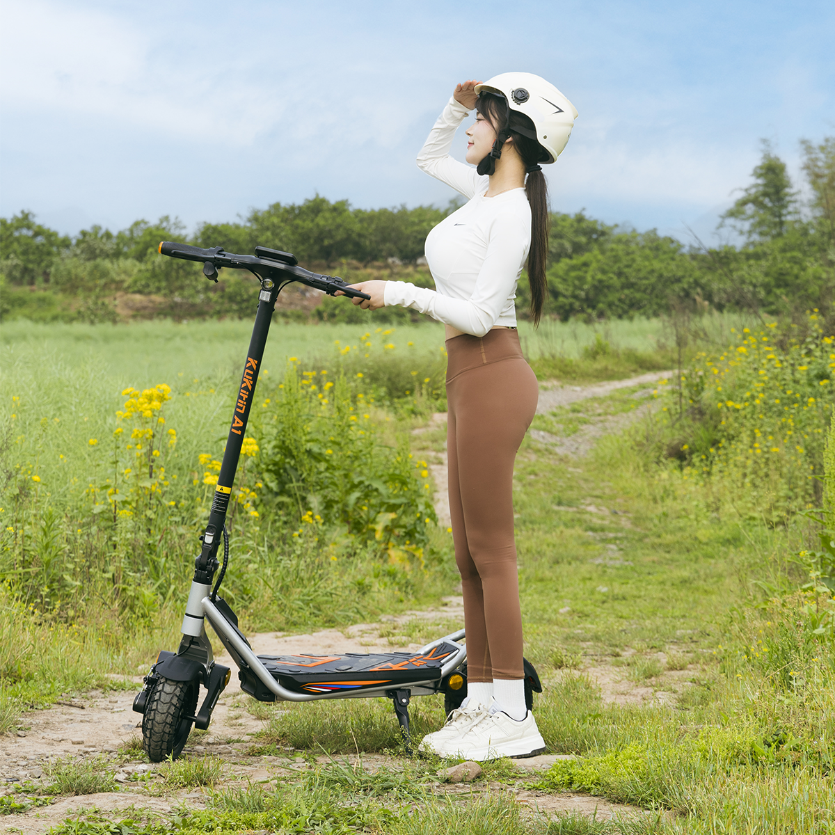 Kobieta w kasku i stroju sportowym stoi obok skutera elektrycznego na trawiastym szlaku.