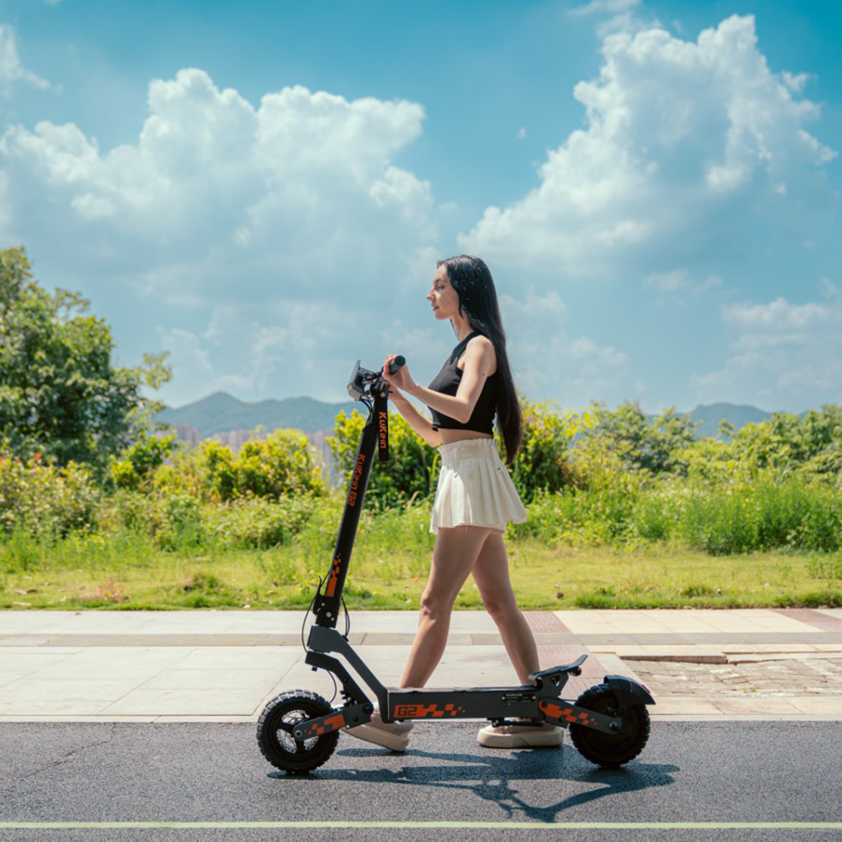 Kobieta idąca z czarno-pomarańczową hulajnogą elektryczną po drodze, niebo i drzewa.