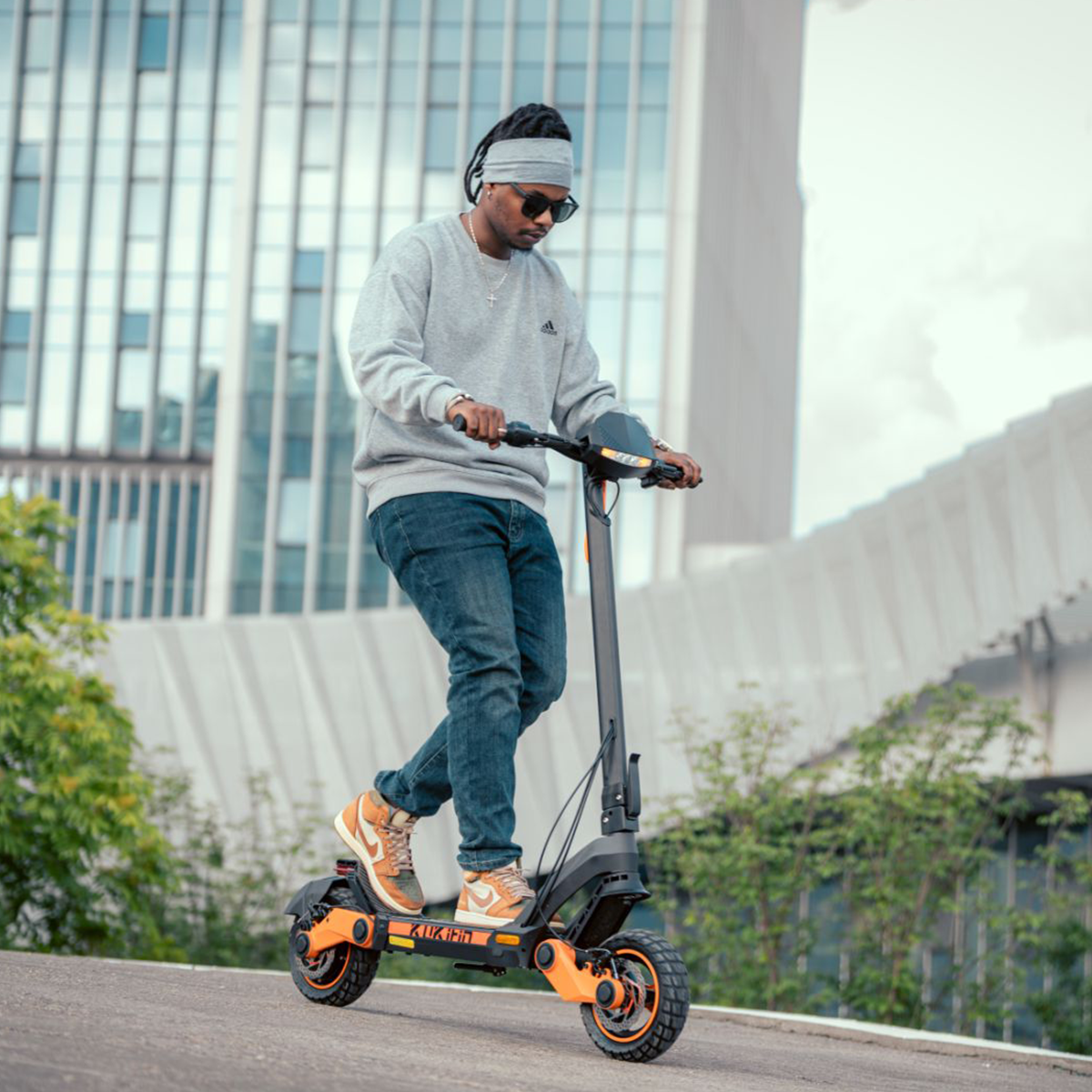 Mężczyzna jadący hulajnogą elektryczną po drodze. Hulajnoga jest pomarańczowo-czarna. Tło to budynek.