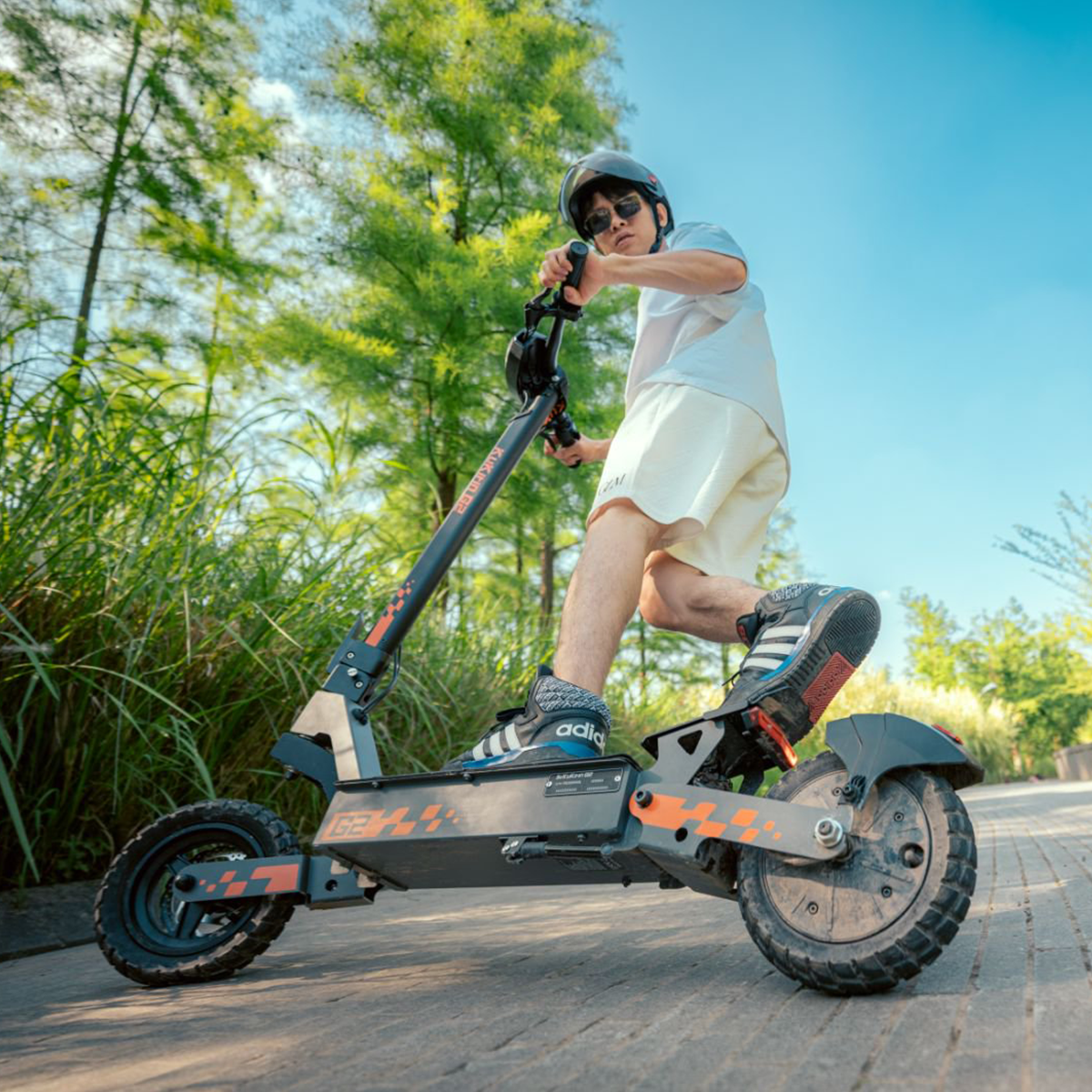 Mężczyzna jadący hulajnogą elektryczną po ścieżce, w kasku i okularach, błękitne niebo.