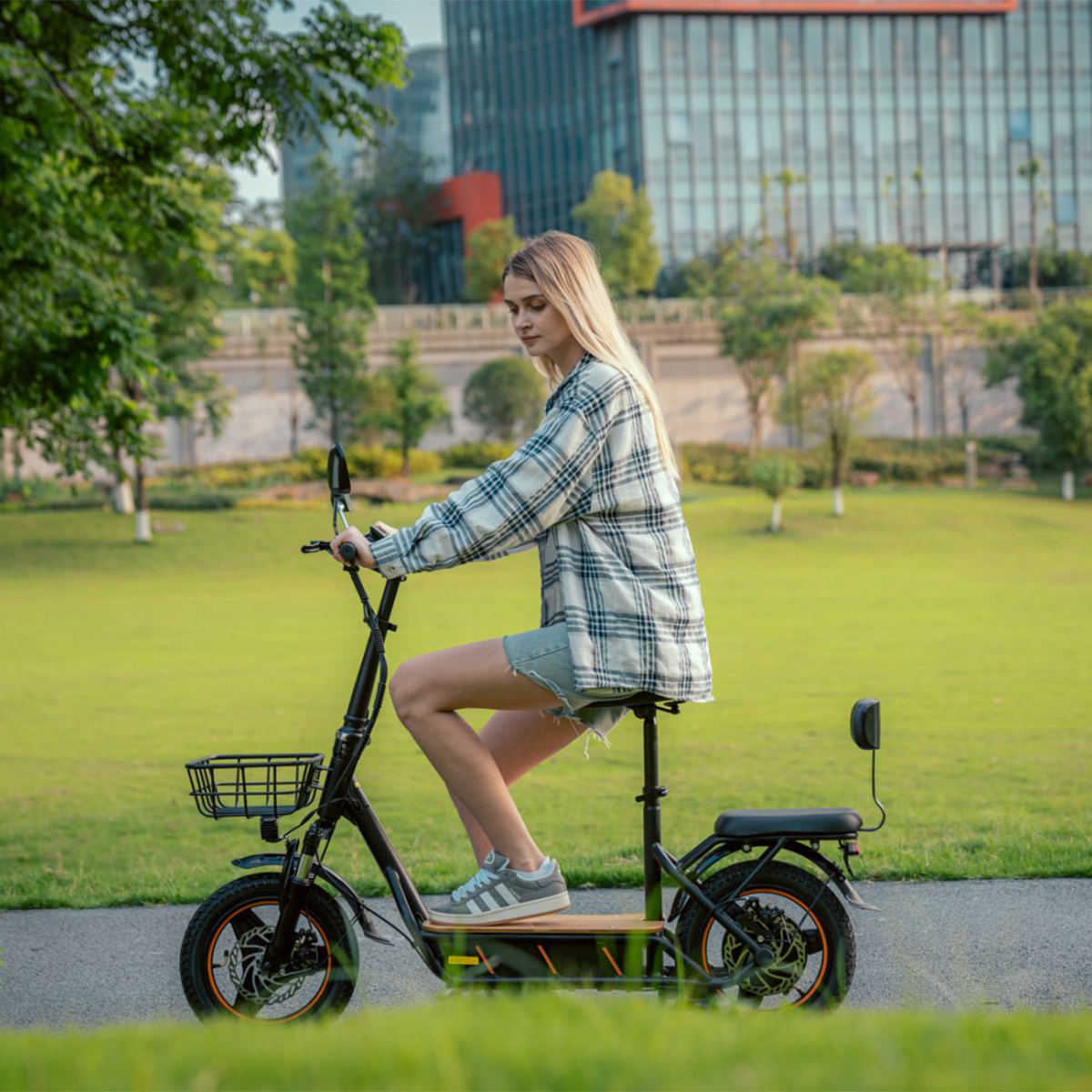 Kobieta jedzie czarną hulajnogą elektryczną po ścieżce. Ma na sobie koszulę w kratę i dżinsowe szorty. Zielona trawa.