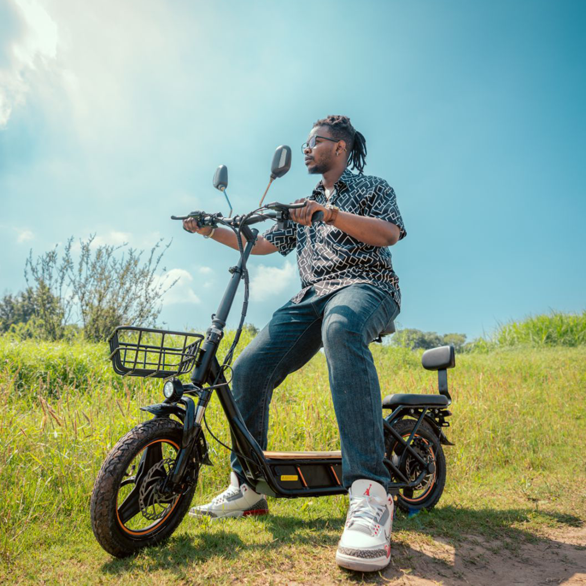 Mężczyzna jedzie hulajnogą elektryczną po trawie. Ma na sobie okulary i koszulę w wzory. Błękitne niebo w tle.