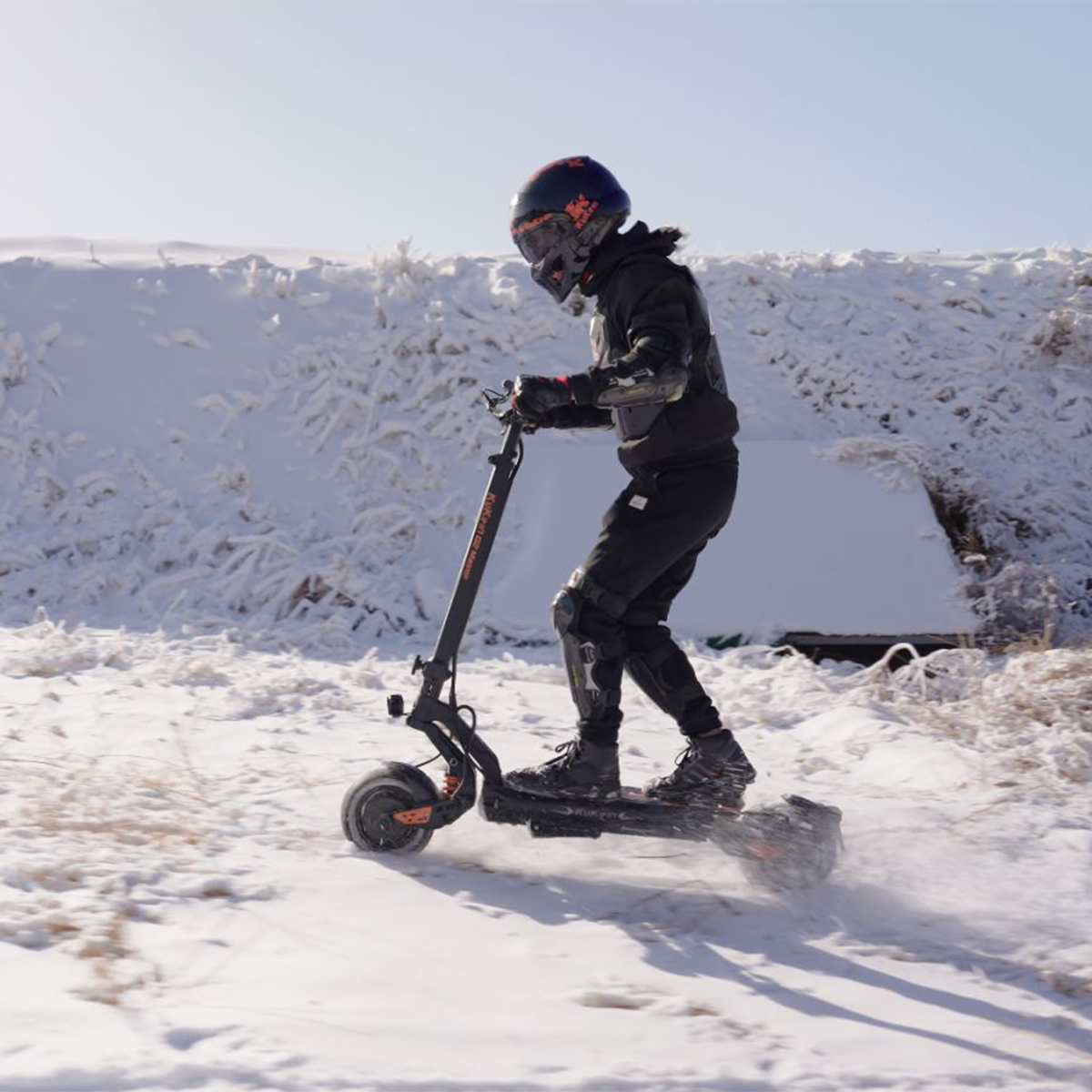 Osoba na hulajnodze elektrycznej na śniegu. Ma kask i ochraniacze. W tle zaśnieżone pole.