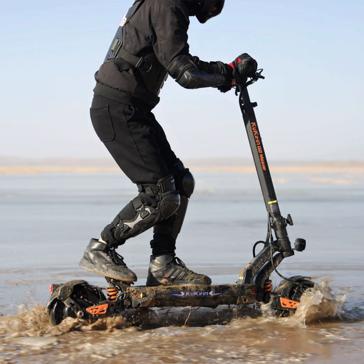Osoba jadąca czarno-pomarańczową hulajnogą elektryczną przez wodę, chlapiąc pojazd. Słonecznie.