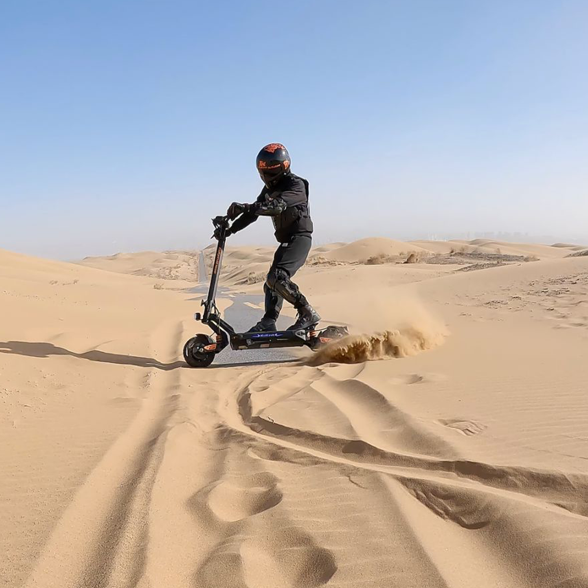 Osoba jadąca czarno-pomarańczową hulajnogą elektryczną na pustyni, wzbijająca piasek. Niebo jest niebieskie.