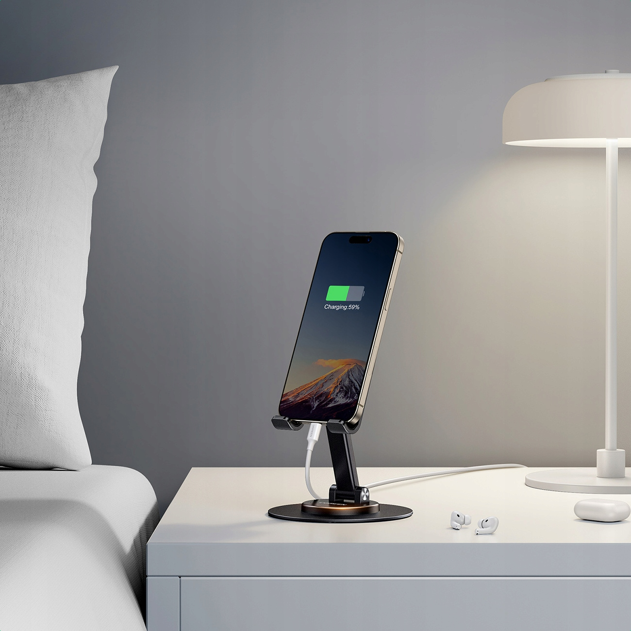 Telefon na stojaku, ładujący się. Biały stół, lampa i poduszka w tle.