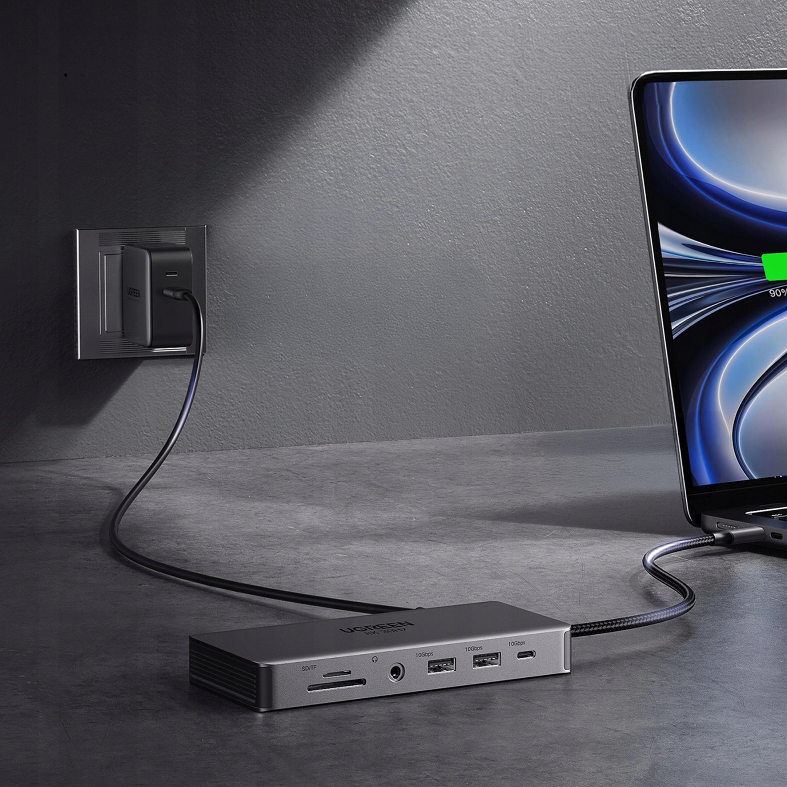 Laptop jest podłączony do stacji dokującej. Zasilacz jest podłączony do ściany. Biurko jest szare.