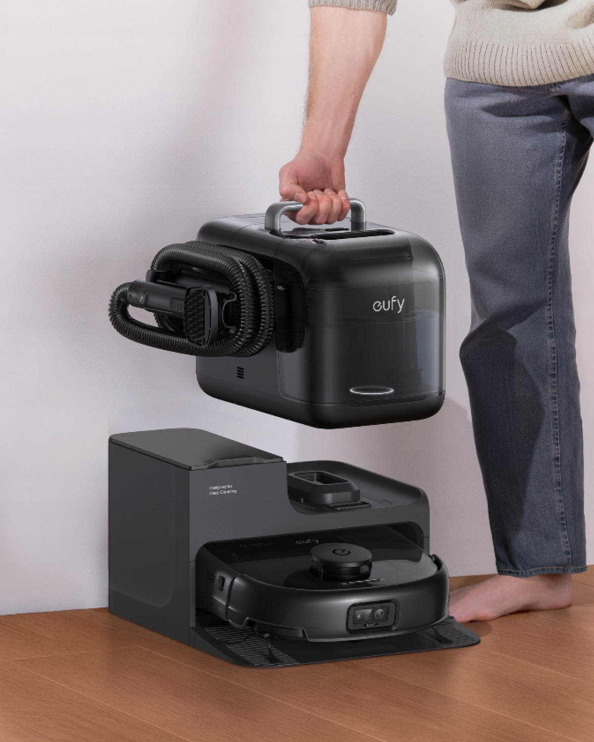 Mężczyzna trzymający czarne urządzenie czyszczące, obok bazy. Baza ma robota sprzątającego. Drewniana podłoga.