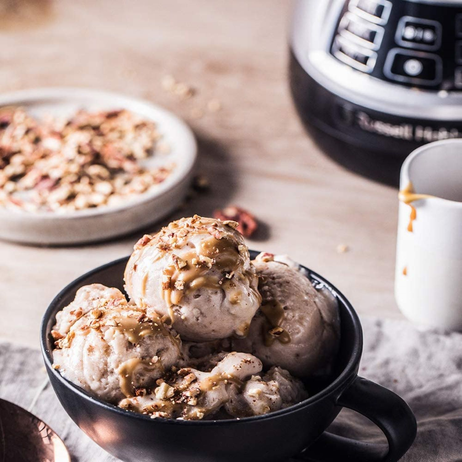Nahaufnahme von Eis in einer schwarzen Schüssel mit Karamell und Nüssen. Im Hintergrund ein Teller mit Nüssen und ein Mixer.