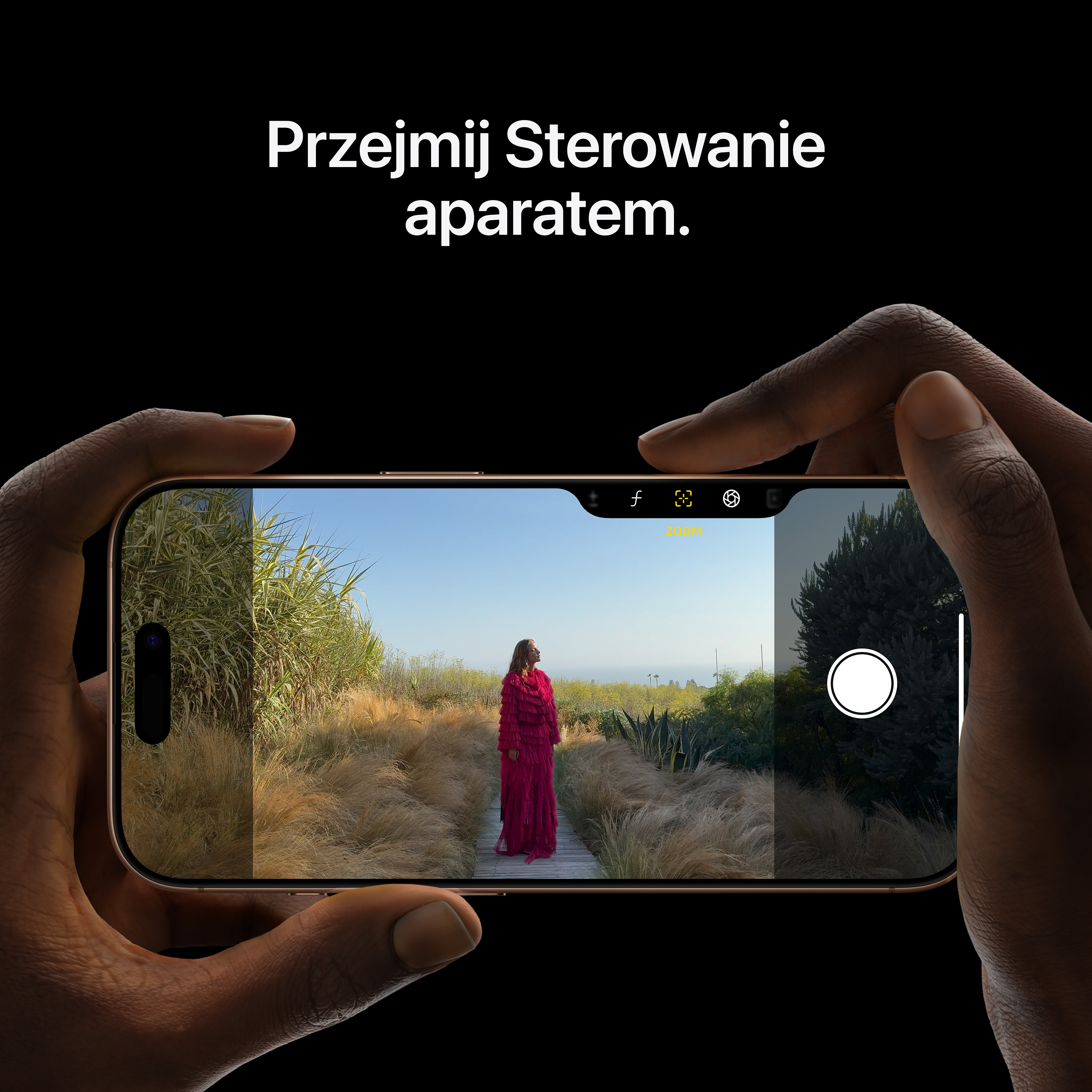 Ręce trzymające telefon, pokazujące kobietę w czerwonej sukience na drewnianej ścieżce, otoczoną zielonymi i żółtymi roślinami.