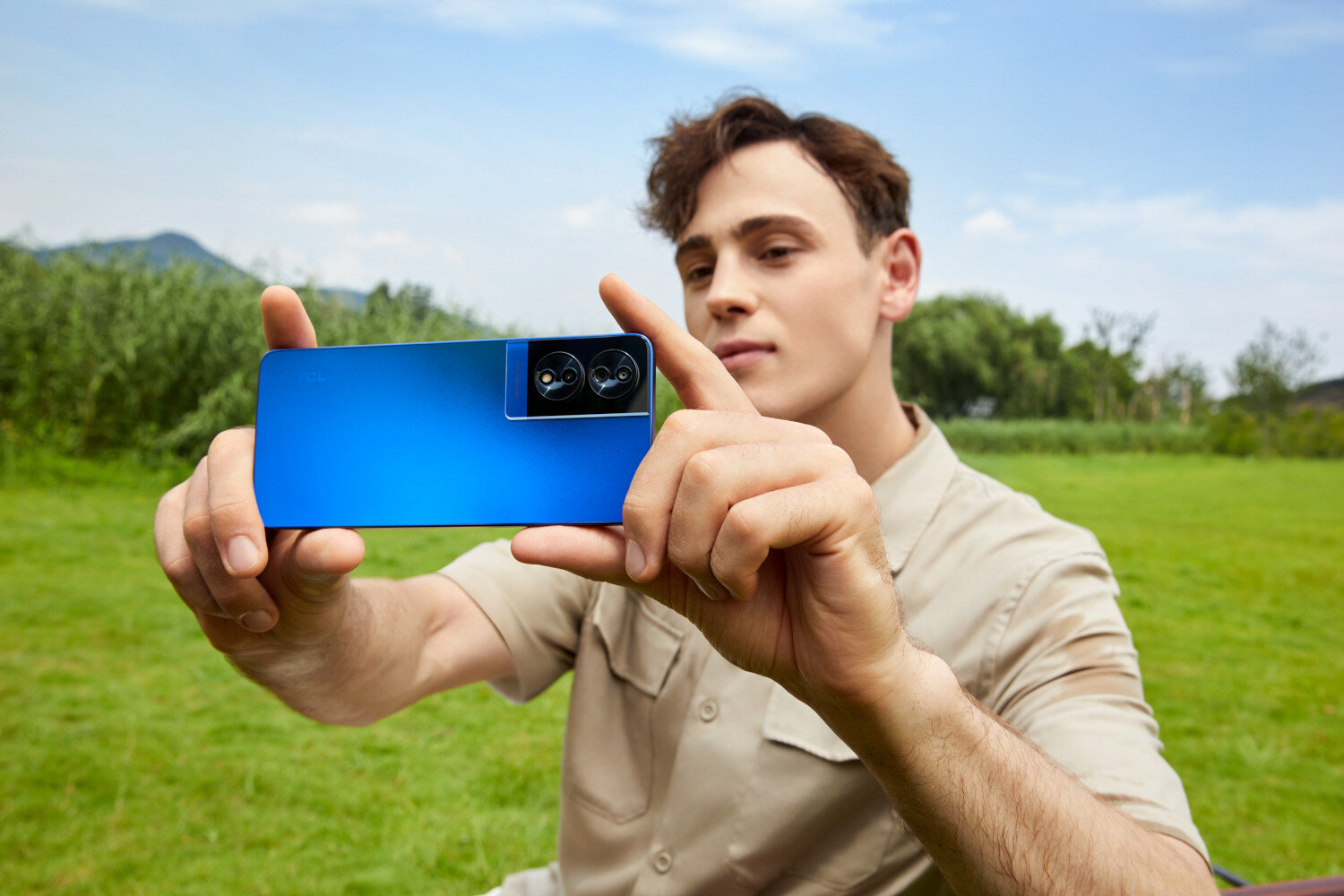 Een man houdt een blauwe telefoon vast en maakt een selfie. Groen gras en bomen op de achtergrond.