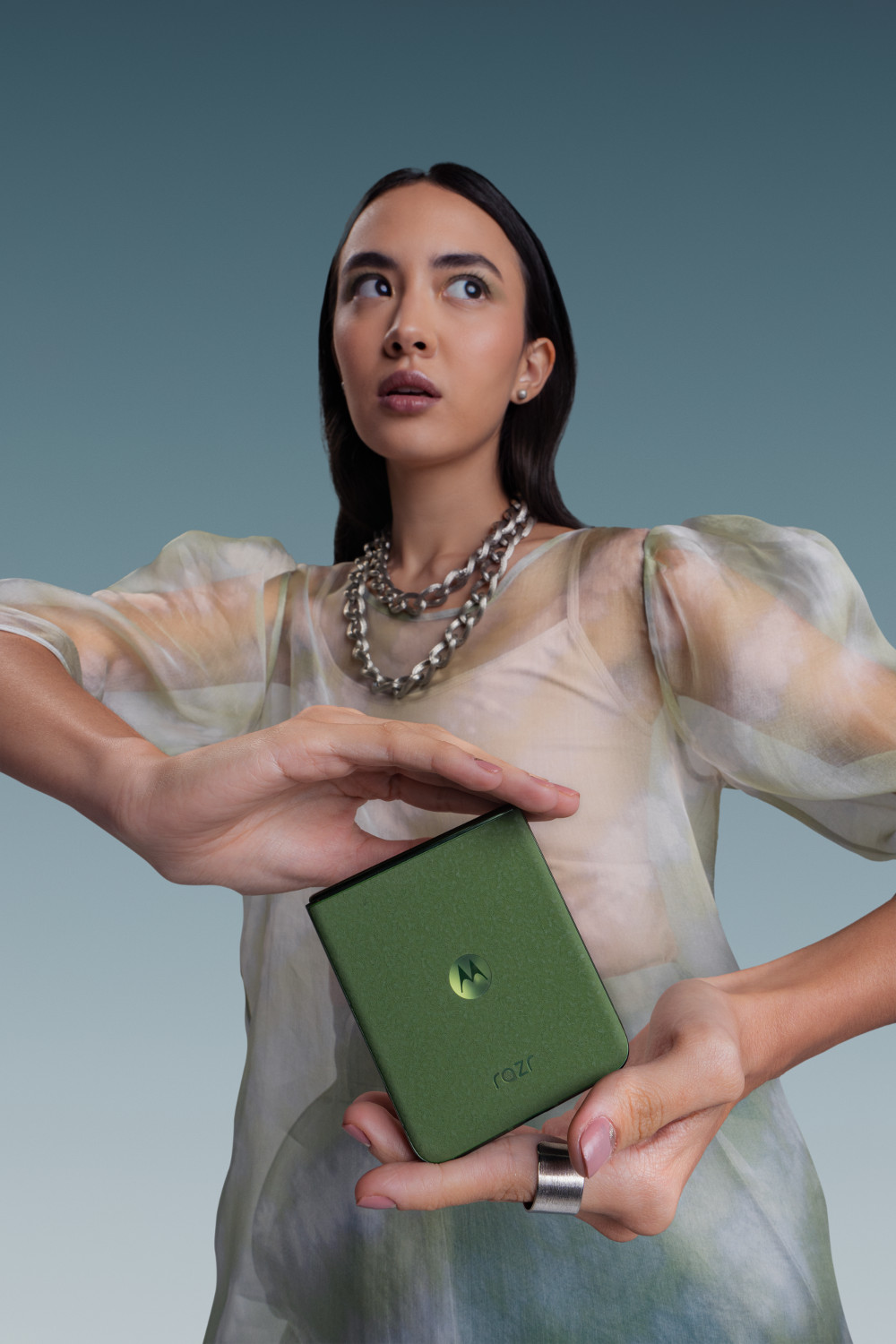 Kobieta trzyma zielony telefon, ma na sobie prześwitującą koszulę we wzory i srebrny naszyjnik.