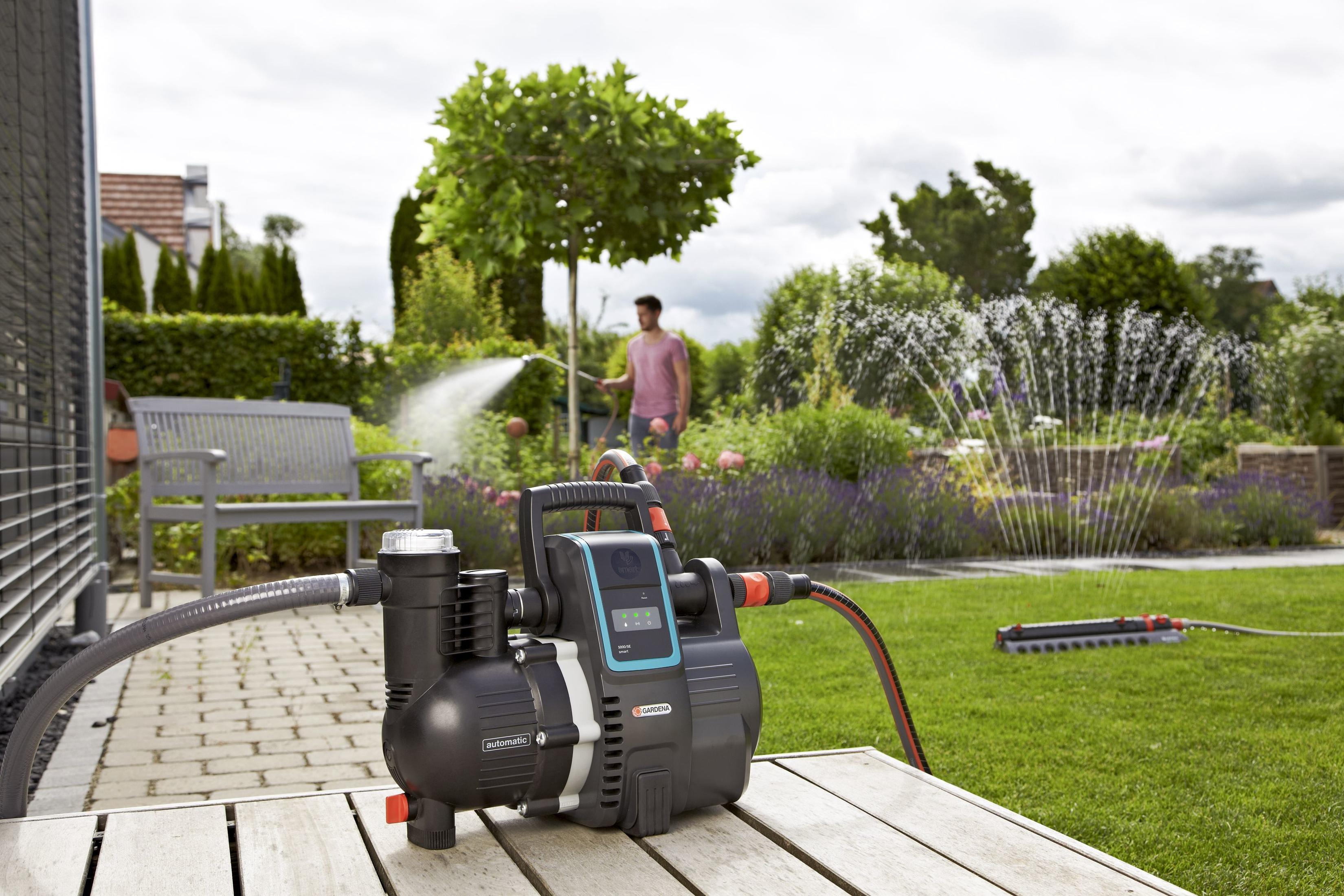Eine schwarze und graue Wasserpumpe auf einem Holzdeck, mit einem Garten im Hintergrund und einem Mann beim Gießen.