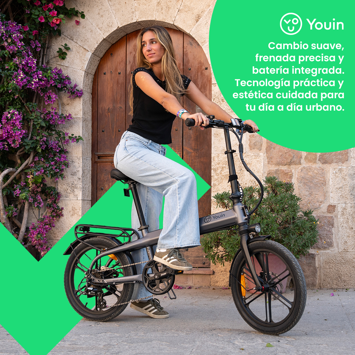 Mujer montando una bicicleta eléctrica gris. Viste vaqueros y top negro. Fondo: puerta.