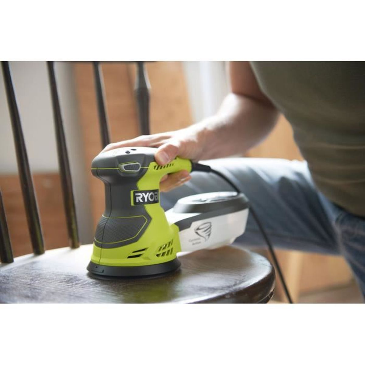 Un uomo che leviga una sedia di legno con una levigatrice Ryobi gialla e nera in un ambiente domestico.
