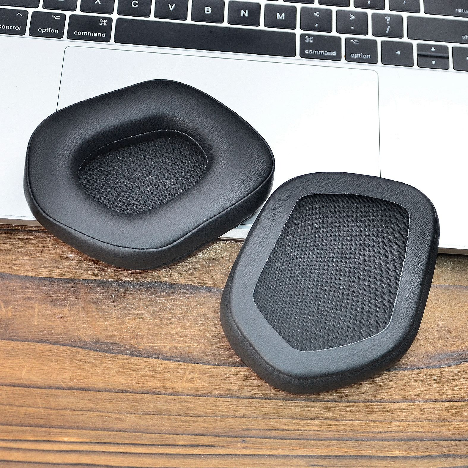 Due cuscinetti auricolari neri per cuffie su una superficie di legno accanto a una tastiera di un laptop.